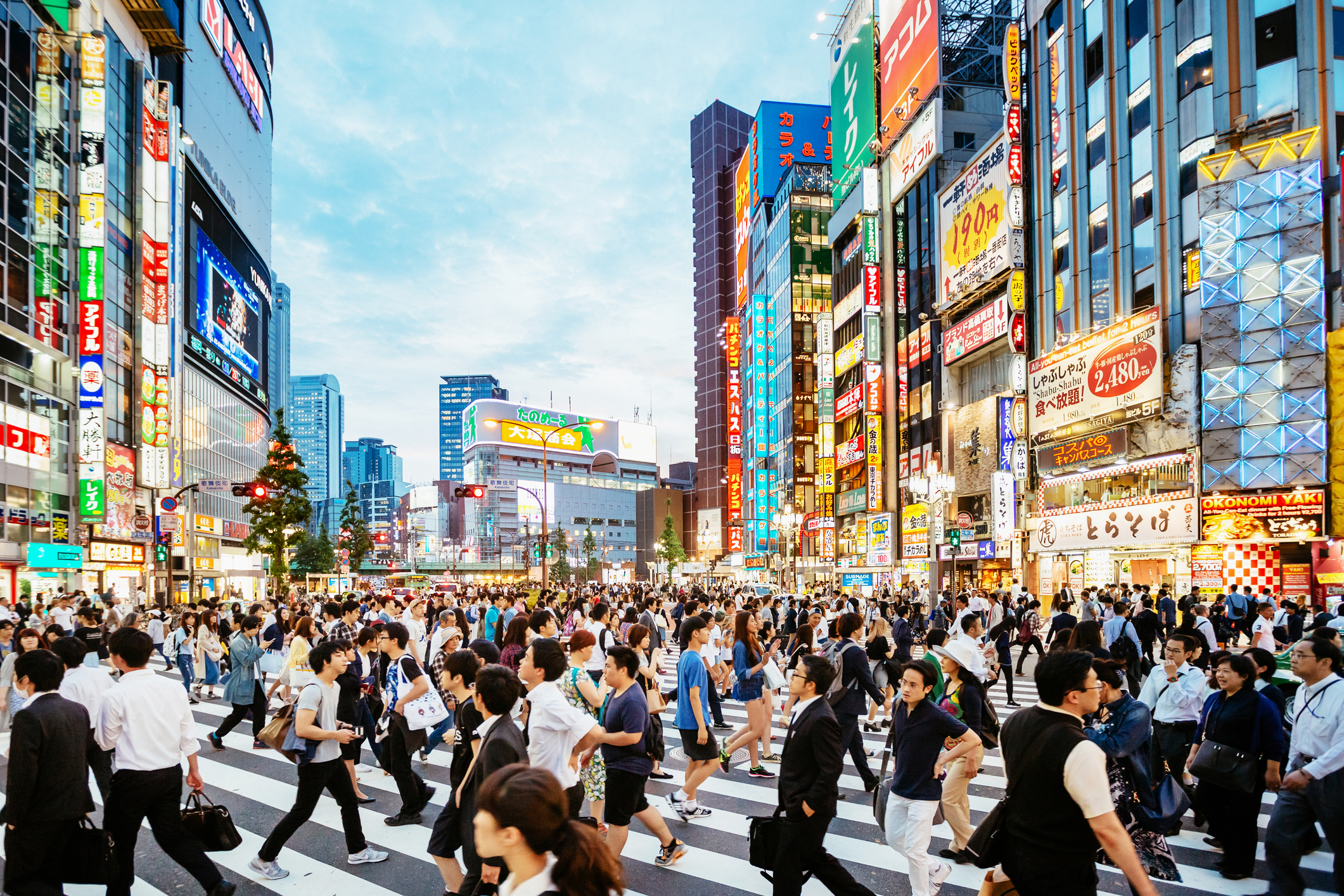 Crowded city street in downtown Tokyo, with people walking in all directions. Bright neon signs and tall buildings fill the background. No celebrities are present