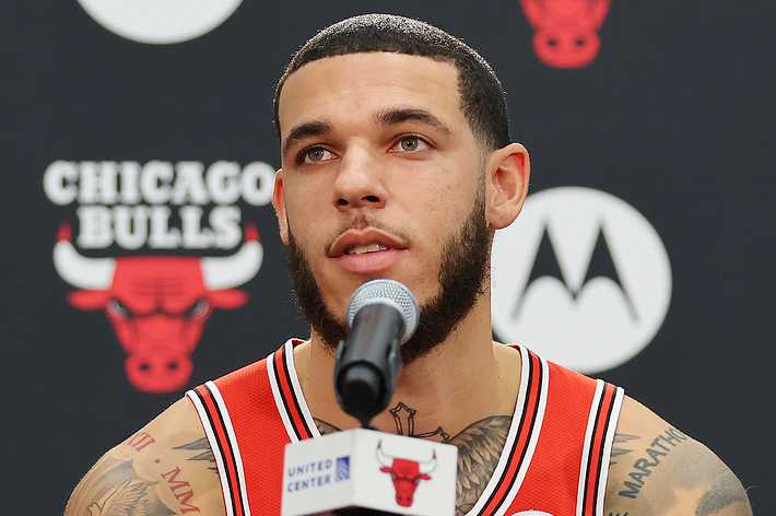 Lonzo Ball speaks at a Chicago Bulls press conference wearing team jersey, microphone in front of him. Bulls logo and sponsor logos are in the background