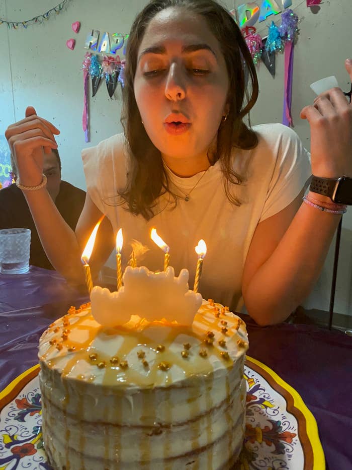 A young woman blows out candles on a birthday cake at a party, with 'Happy Birthday' decorations in the background