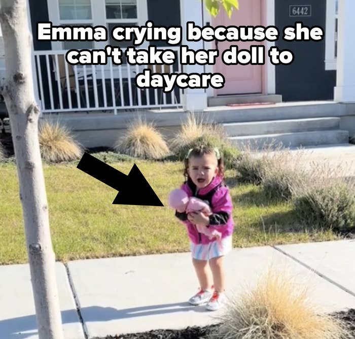 A young girl stands on a sidewalk crying and holding a doll with a house and garden in the background