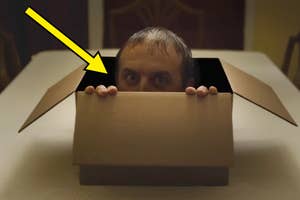 A person is hiding inside a partially opened cardboard box on a table, with only their head and hands visible. Yellow arrow points to their face