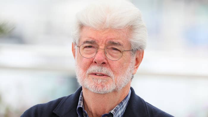 George Lucas, wearing glasses and a beard, looks thoughtful in a casual jacket and checkered shirt