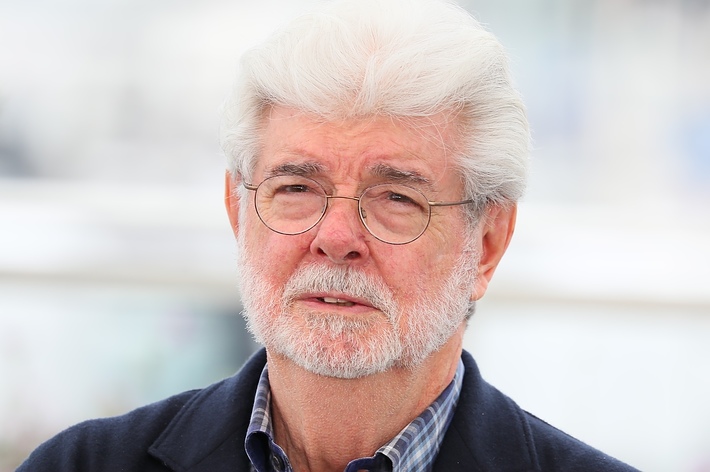 An older man with glasses and a beard is wearing a dark jacket and a checkered shirt, looking into the distance