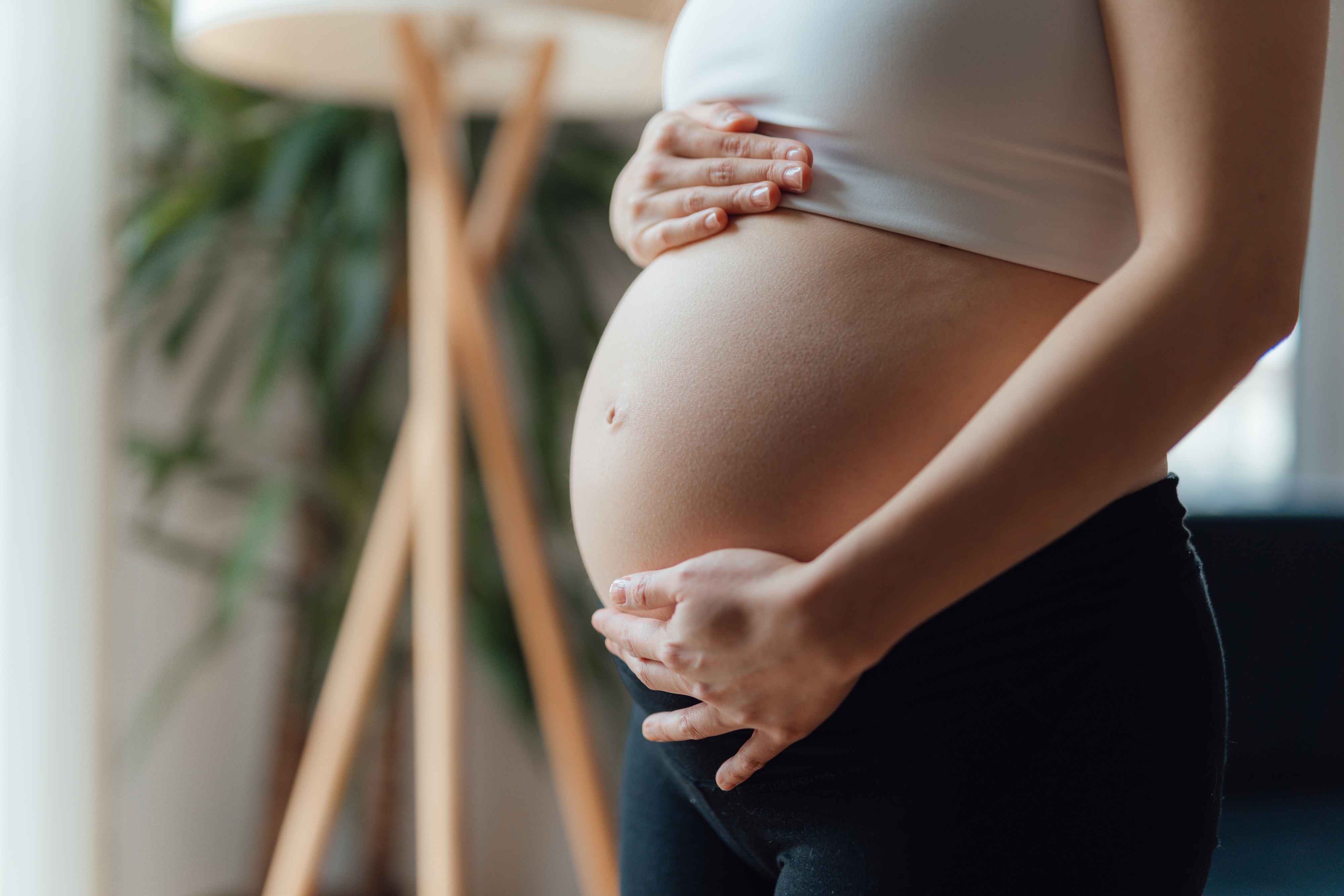 A person in a sleeveless top holds their pregnant belly. A plant and floor lamp are in the background