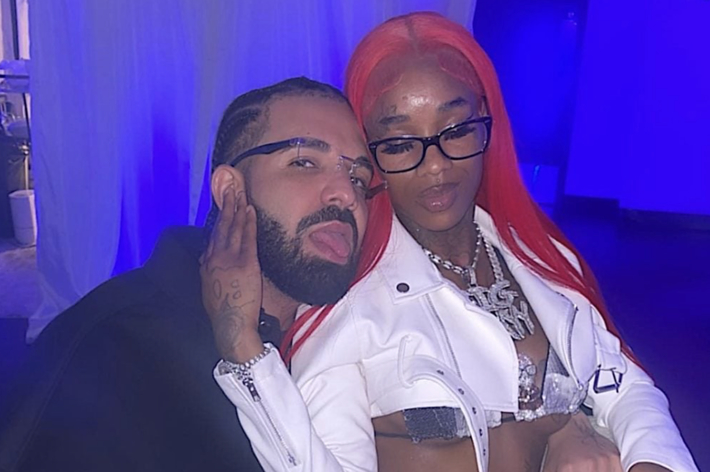 Drake and Sexyy Red pose together, smiling. Sexyy Red wears a silver and white outfit with a white jacket and glasses, both adorned with jewelry