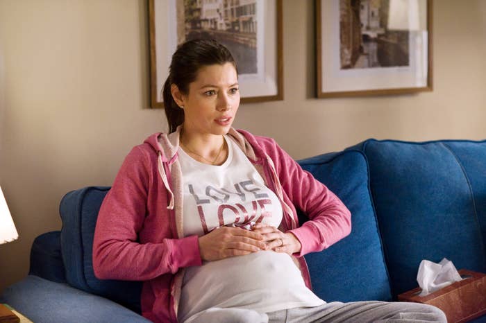 Jessica Biel, in casual clothing, sits on a couch with her hands on her pregnant belly, wearing a "Love" t-shirt