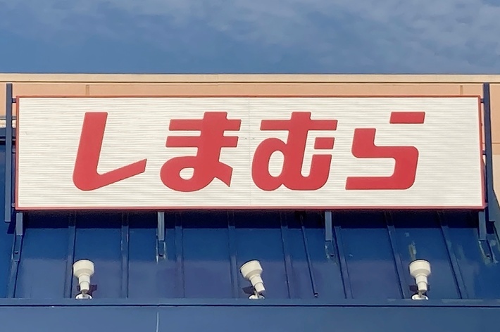 Storefront with a sign in Japanese that reads “Shimamura” above an entrance with blue panels. There is a handicap parking sign visible in the foreground