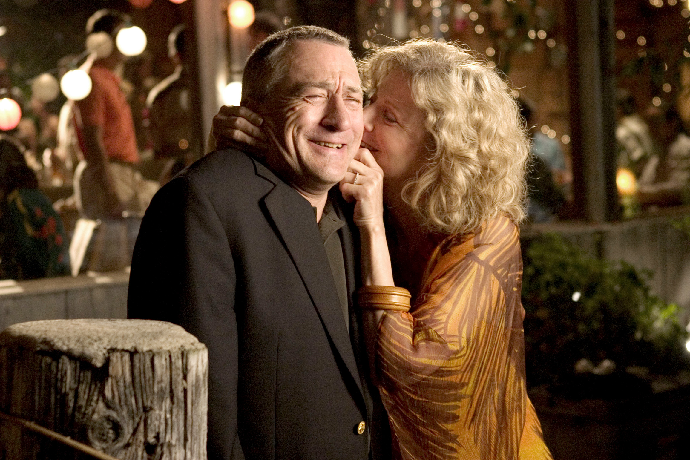 Robert De Niro and Blythe Danner share a laugh; Danner whispers in De Niro's ear while holding his face playfully, both dressed in smart-casual attire