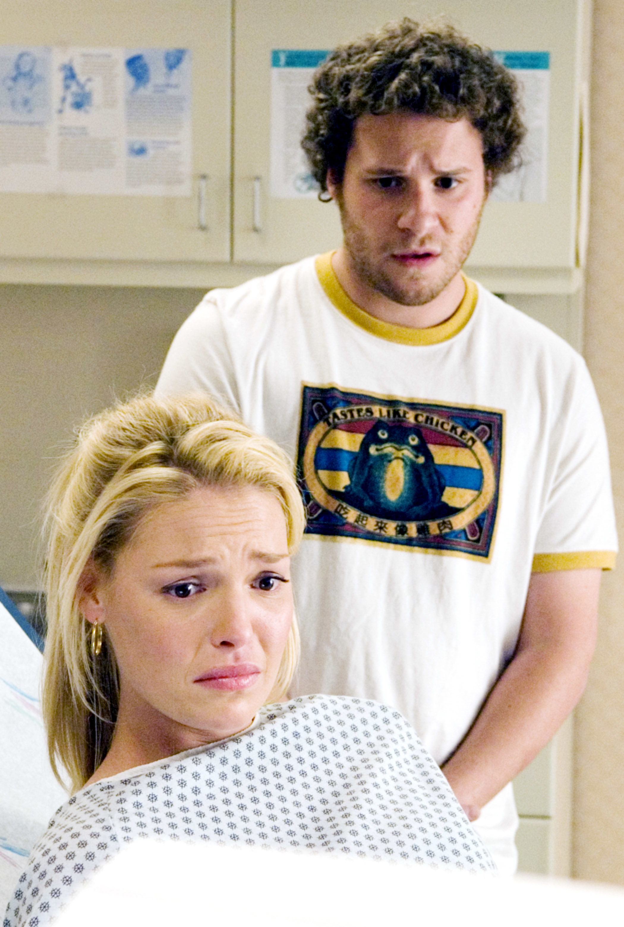 Katherine Heigl sits distressed in a hospital gown while Seth Rogen, wearing a graphic T-shirt, stands behind her looking concerned