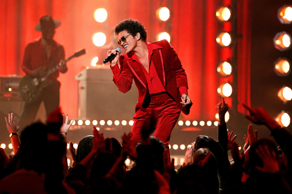 Bruno Mars performs on stage in a stylish suit, singing into a microphone, with an enthusiastic crowd and backup band