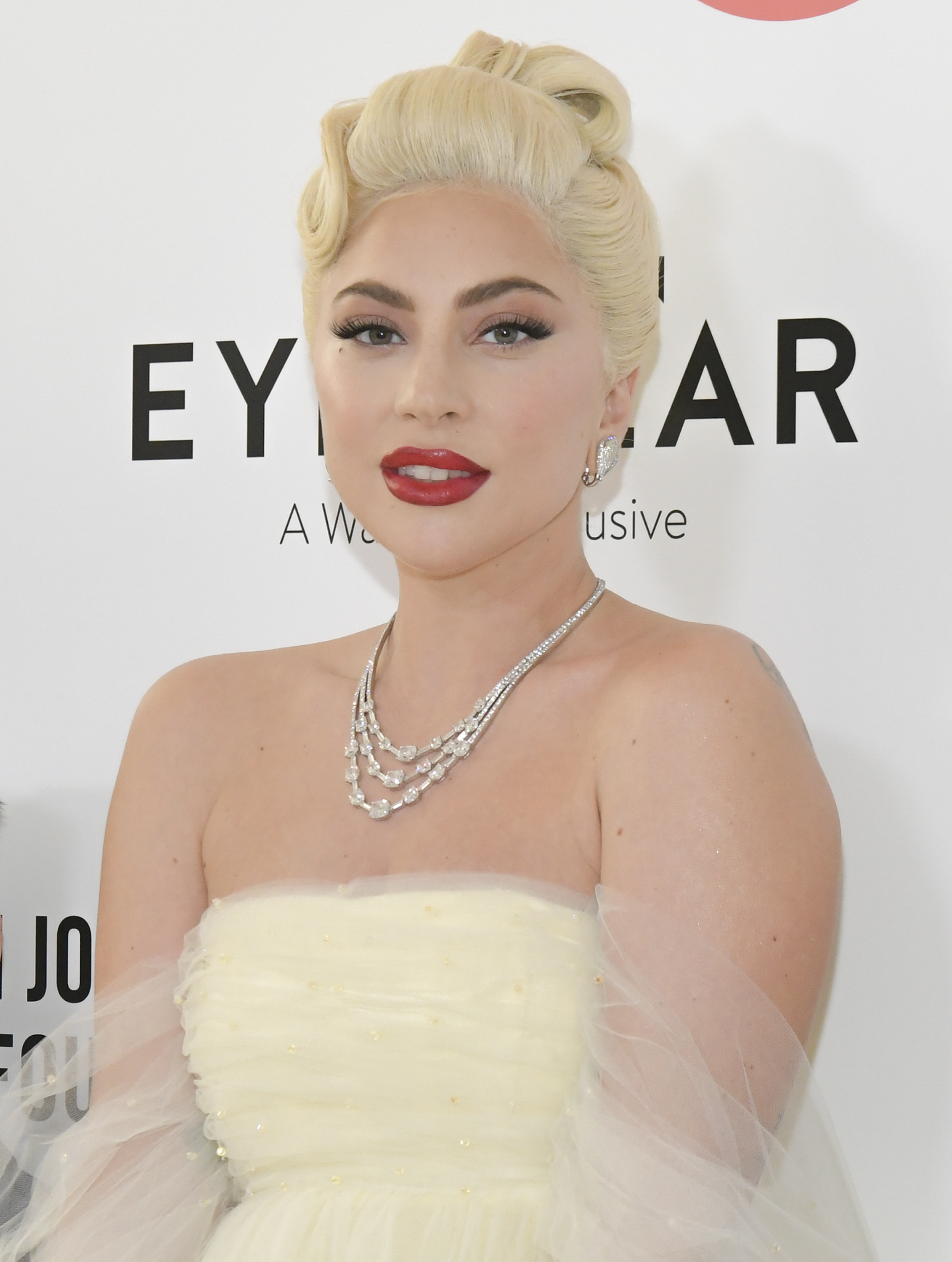 Lady Gaga on the red carpet, wearing an elegant strapless gown and a layered diamond necklace, with retro-styled hair and bold makeup