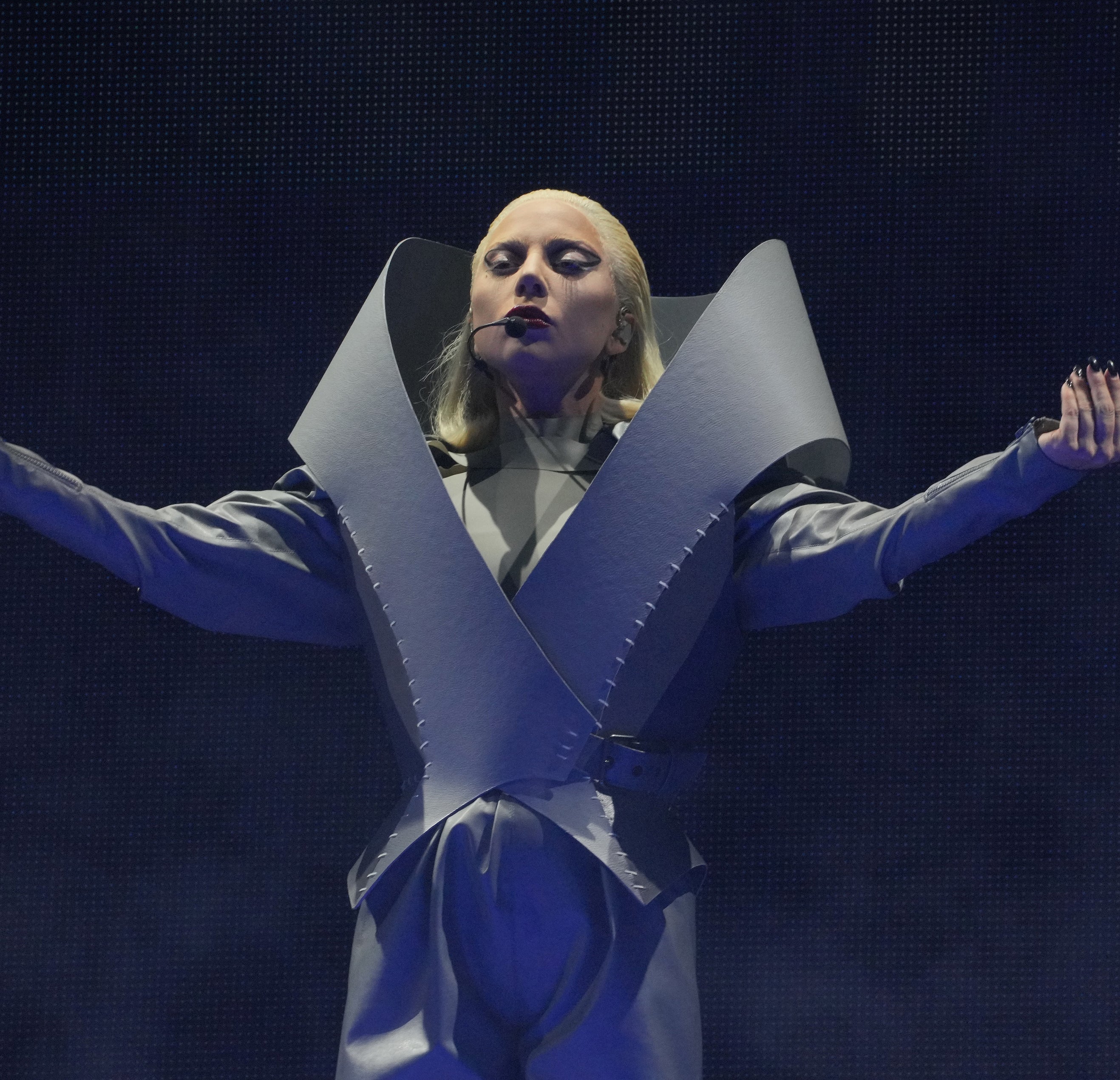 Lady Gaga is on stage, wearing an avant-garde, structured outfit with dramatic, oversized shoulders and striking poses during a performance