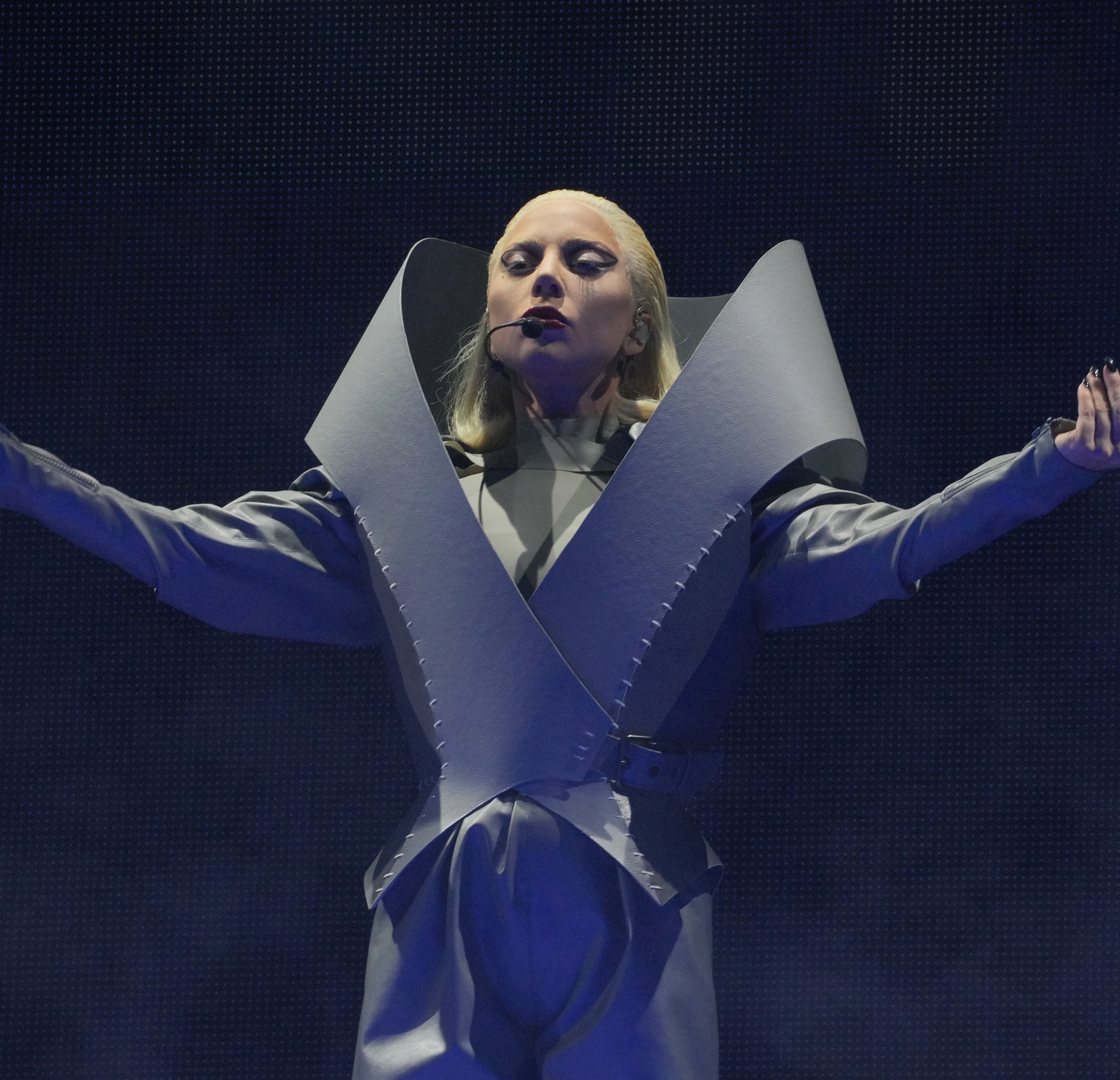 Lady Gaga is on stage, wearing an avant-garde, structured outfit with dramatic, oversized shoulders and striking poses during a performance