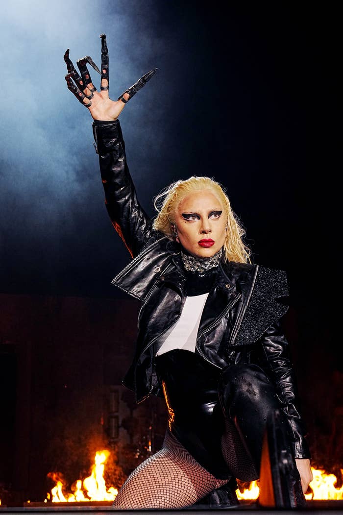 Lady Gaga performing on stage, wearing a leather jacket, pants, fishnet stockings, and holding up a clawed glove with flames in the background