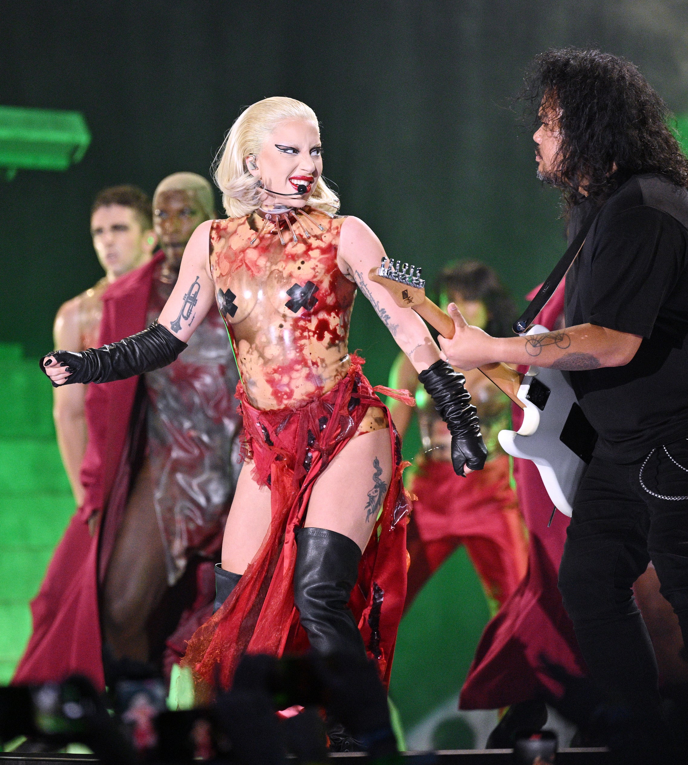 Lady Gaga, in a unique, avant-garde dress, performs with guitarist on stage, surrounded by backup dancers in futuristic attire