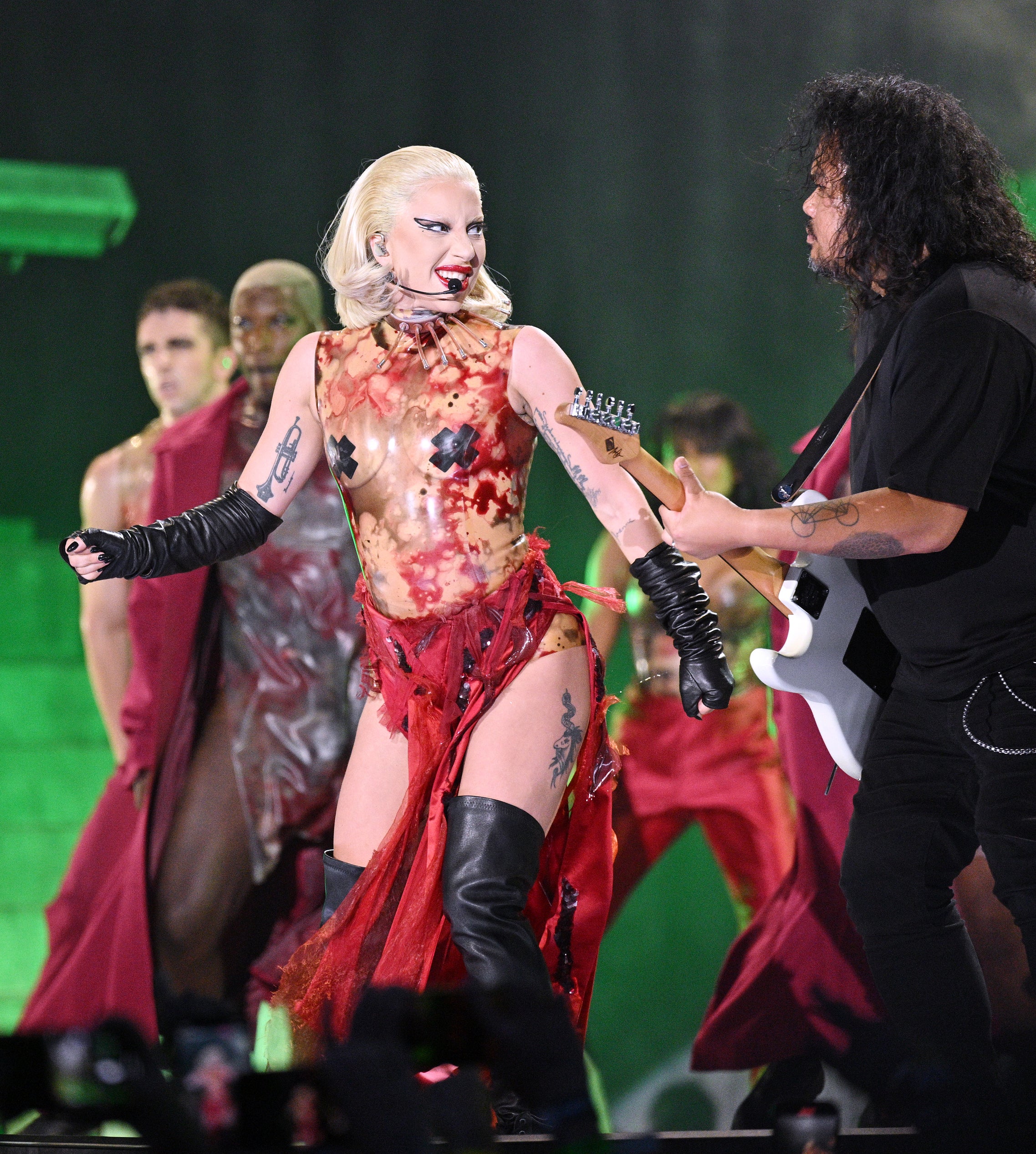 Lady Gaga, in a unique, avant-garde dress, performs with guitarist on stage, surrounded by backup dancers in futuristic attire