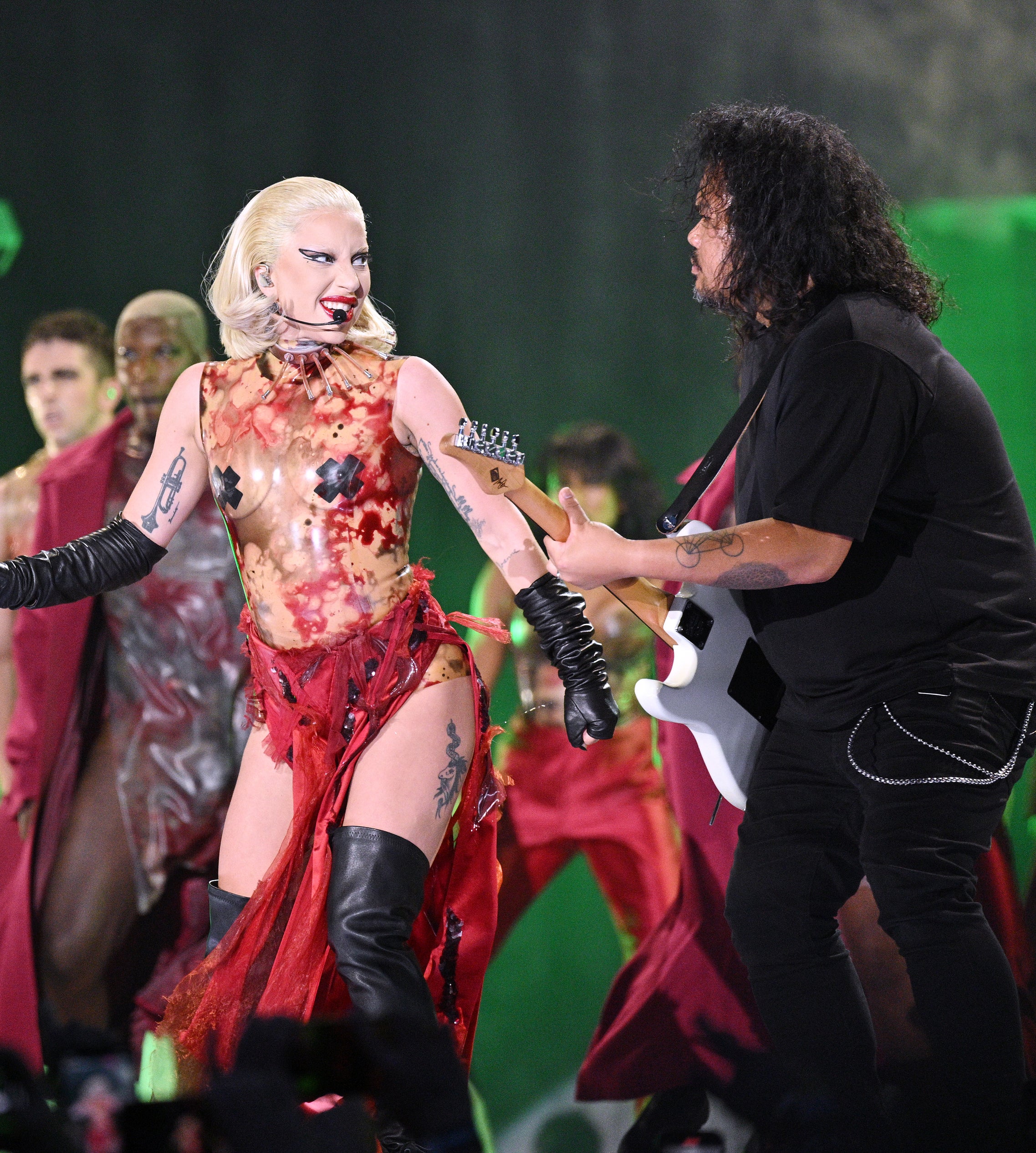 Lady Gaga, in a unique, avant-garde dress, performs with guitarist on stage, surrounded by backup dancers in futuristic attire