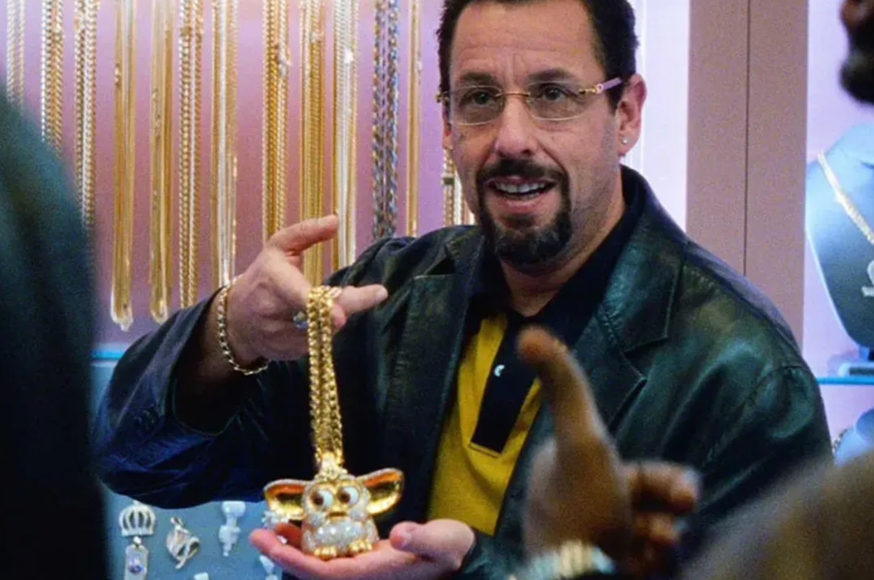 Adam Sandler, in a casual outfit with a leather jacket, holds up a gold necklace with a pendant resembling Gizmo from the movie "Gremlins."
