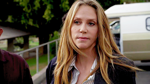 A woman with long blonde hair, wearing a striped shirt and a blazer, stands outside near a van, looking serious