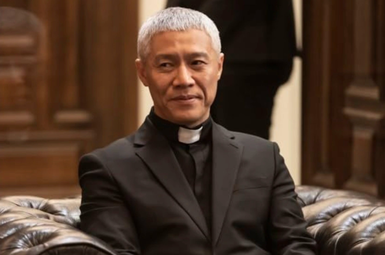 Andrew Koji is seated wearing a formal black priest uniform in a room with wooden decor