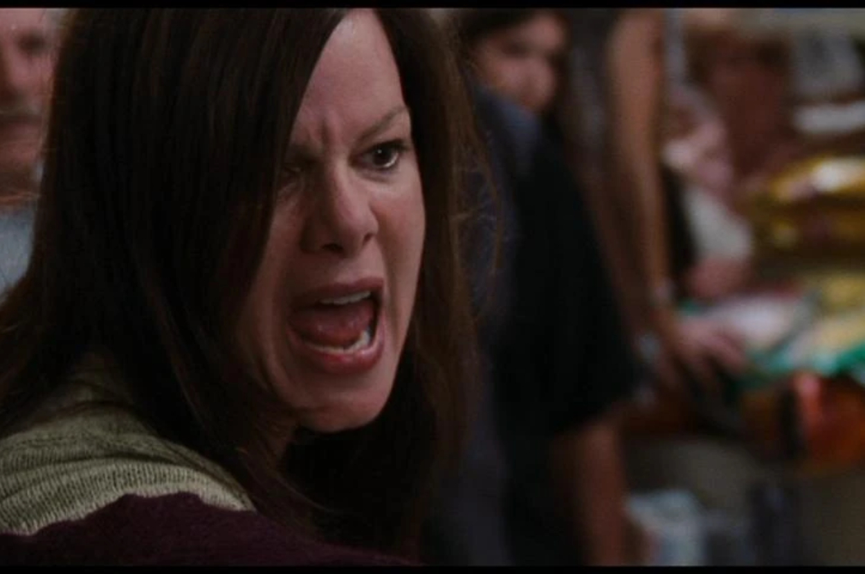 Woman with long hair yelling, close-up. Background shows a crowded room, likely indicating a tense or dramatic situation. No additional context or other identifiable persons