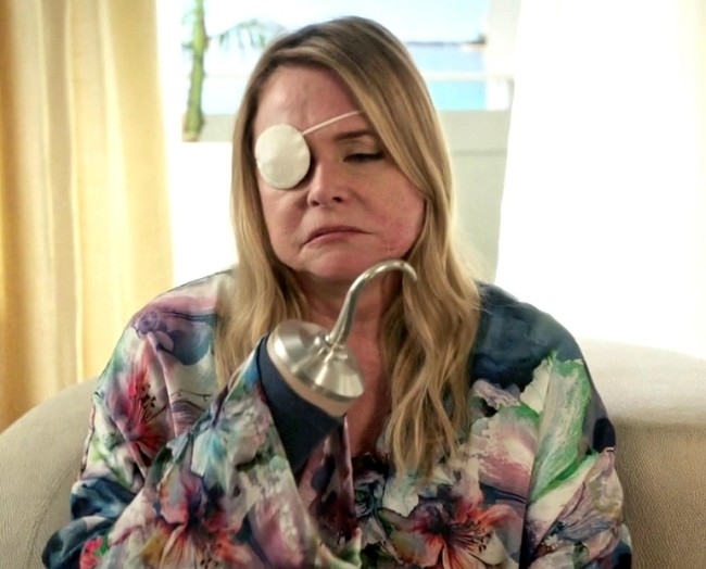 Jennifer Coolidge with an eye patch and a hook hand prop in a floral robe, seated indoors