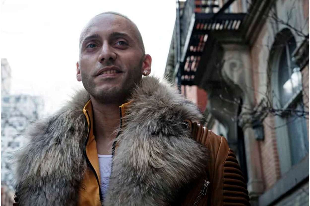 Person wearing a fur-collared jacket, looking off camera on a city street with brick buildings in the background