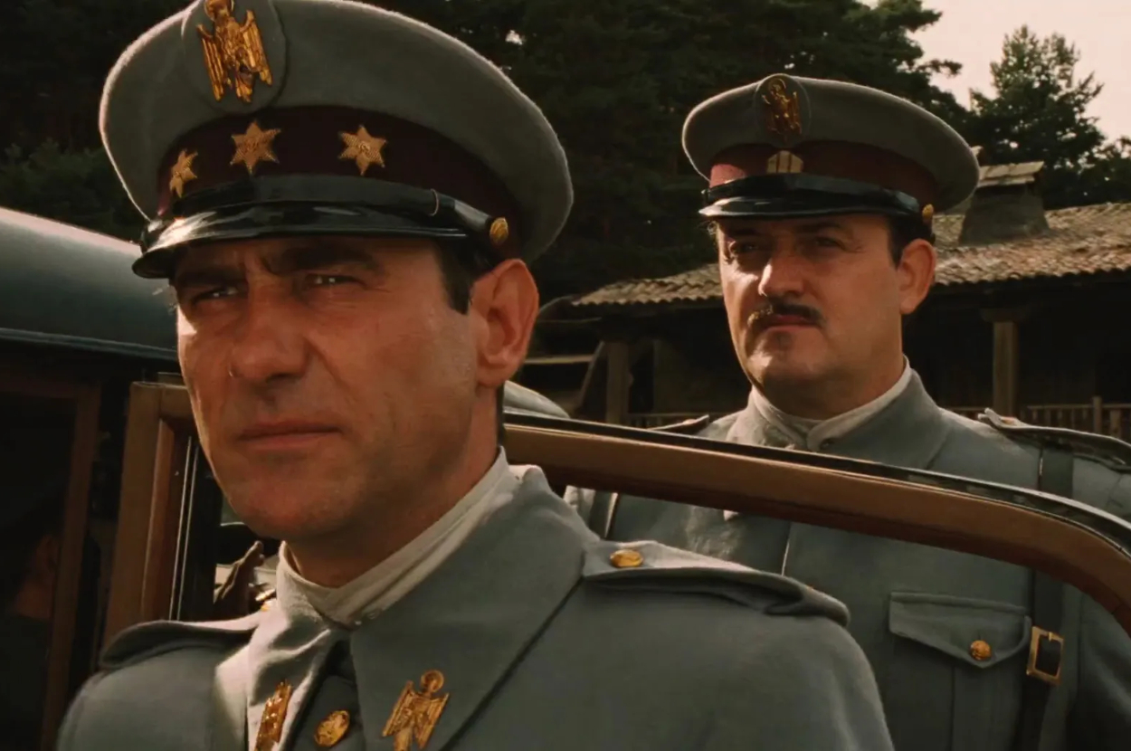 Two men in military uniforms, one closer to the camera with a serious expression and the other behind him, in a scene from a movie