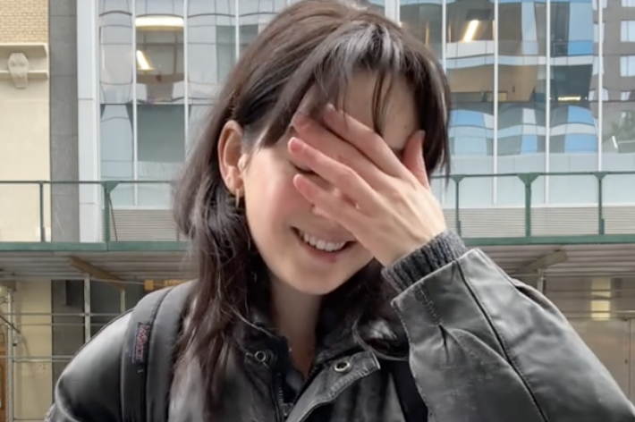 女性が笑顔で紙を持ちながら目を覆っています。街中での様子です。人の名前はわかりません。