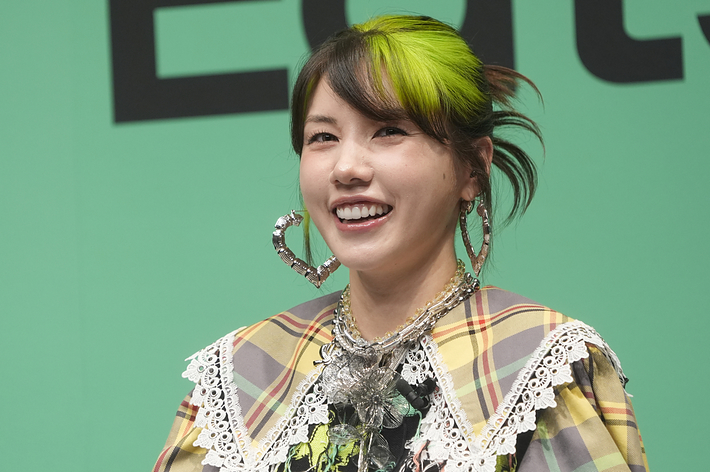 アイドルの篠崎愛がイベントで笑顔を見せています。彼女はチェック柄の衣装に大きなハートのイヤリングと独特なネックレスを着用しています。