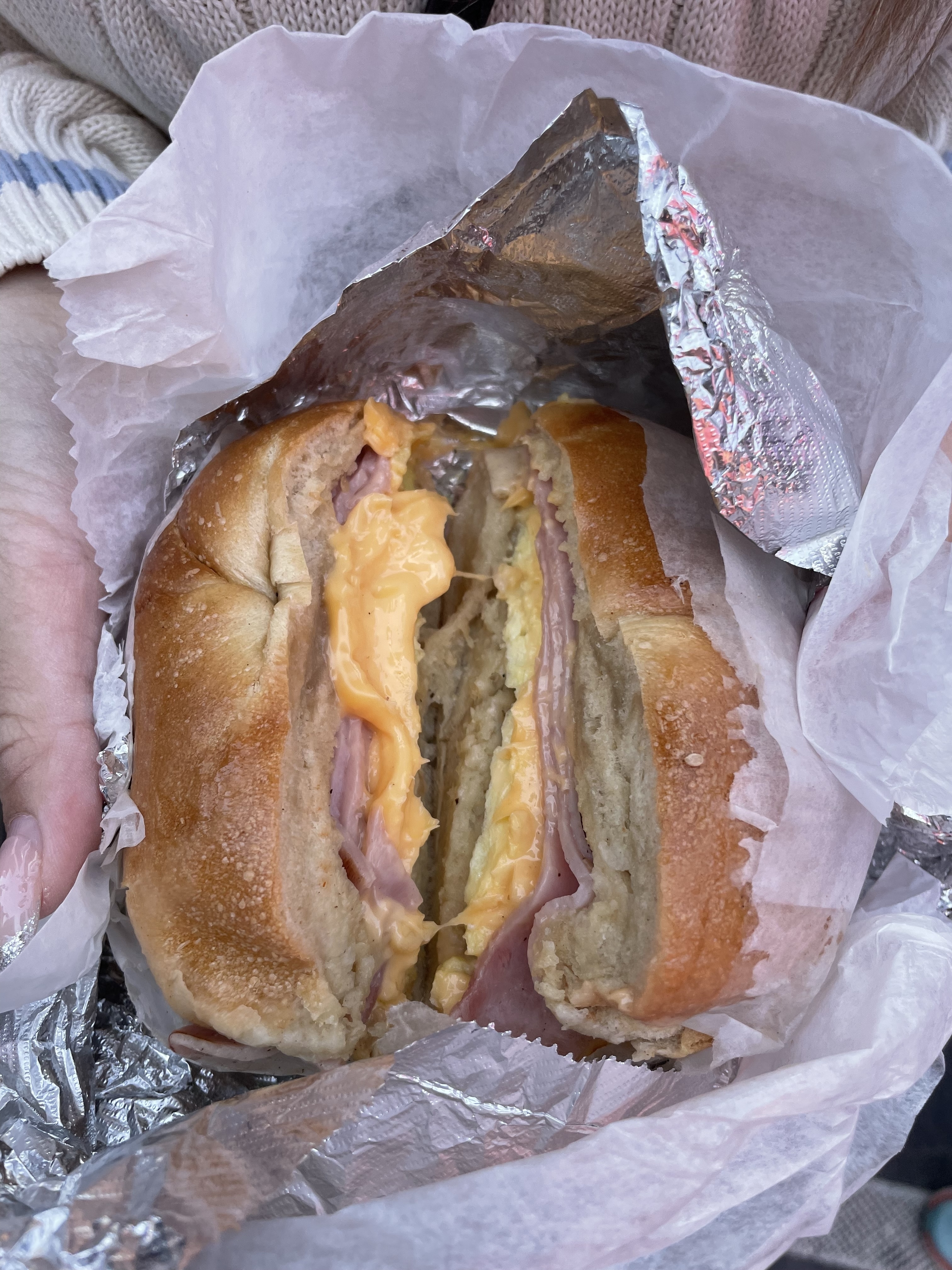 Close-up of a breakfast sandwich with ham, scrambled eggs, and melted cheese in a bagel, held in a hand with a foil wrapper partially removed