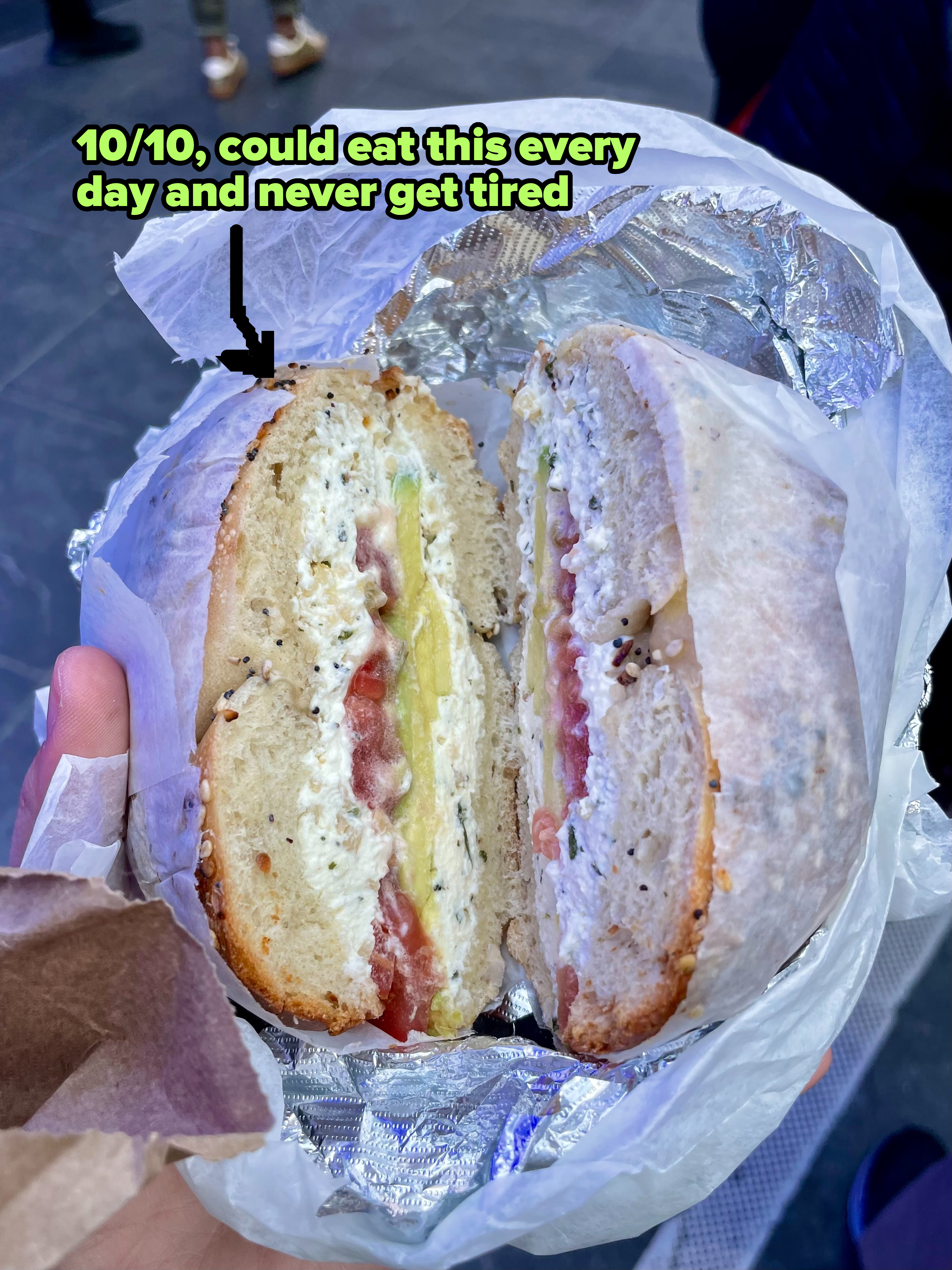 A person holds a bagel sandwich cut in half, filled with cream cheese, tomato, and cucumber, wrapped in foil and parchment paper