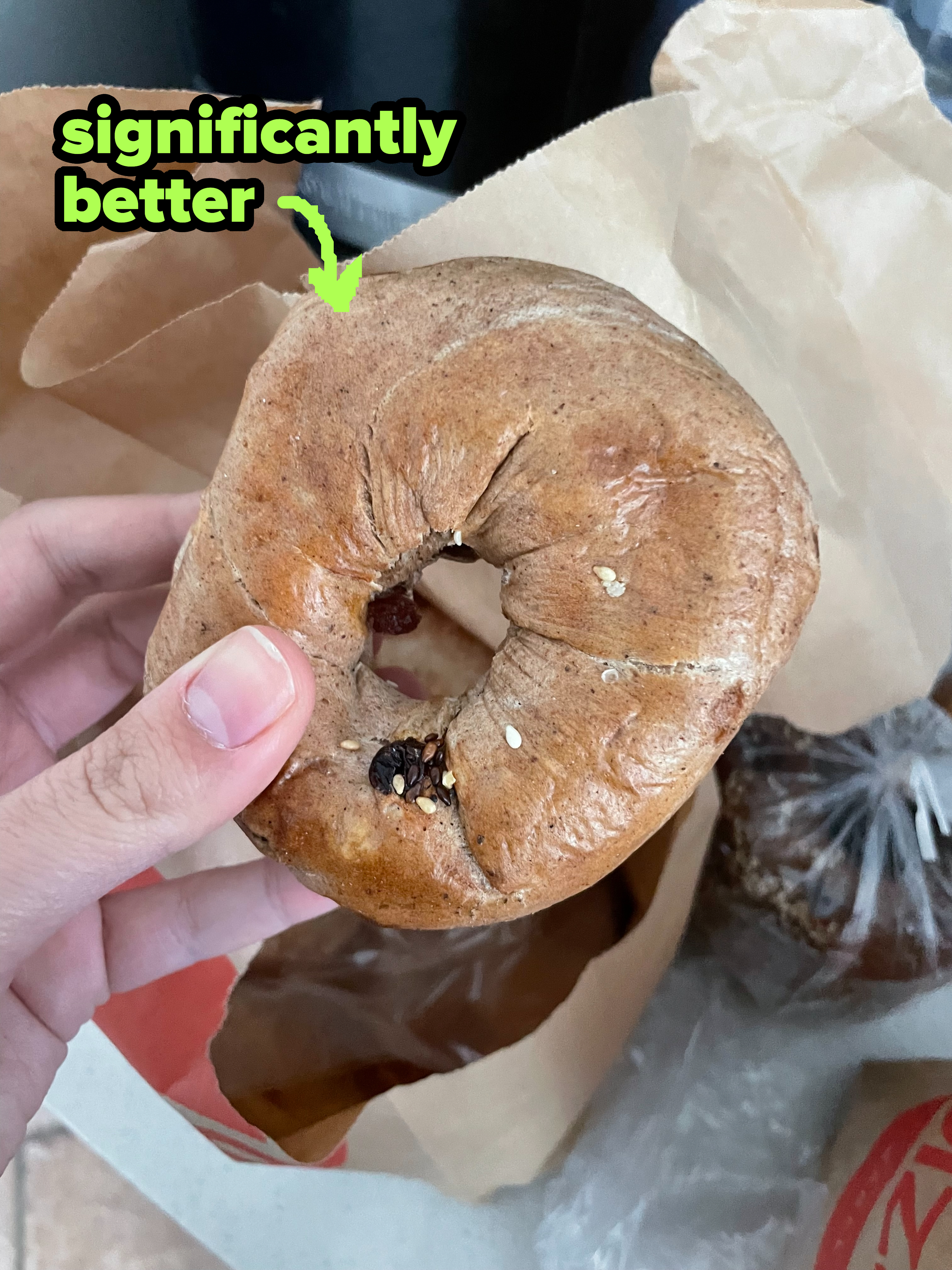 A hand holding a bagel with visible sesame seeds and what appears to be raisins or currants embedded in it. Background features a brown paper bag