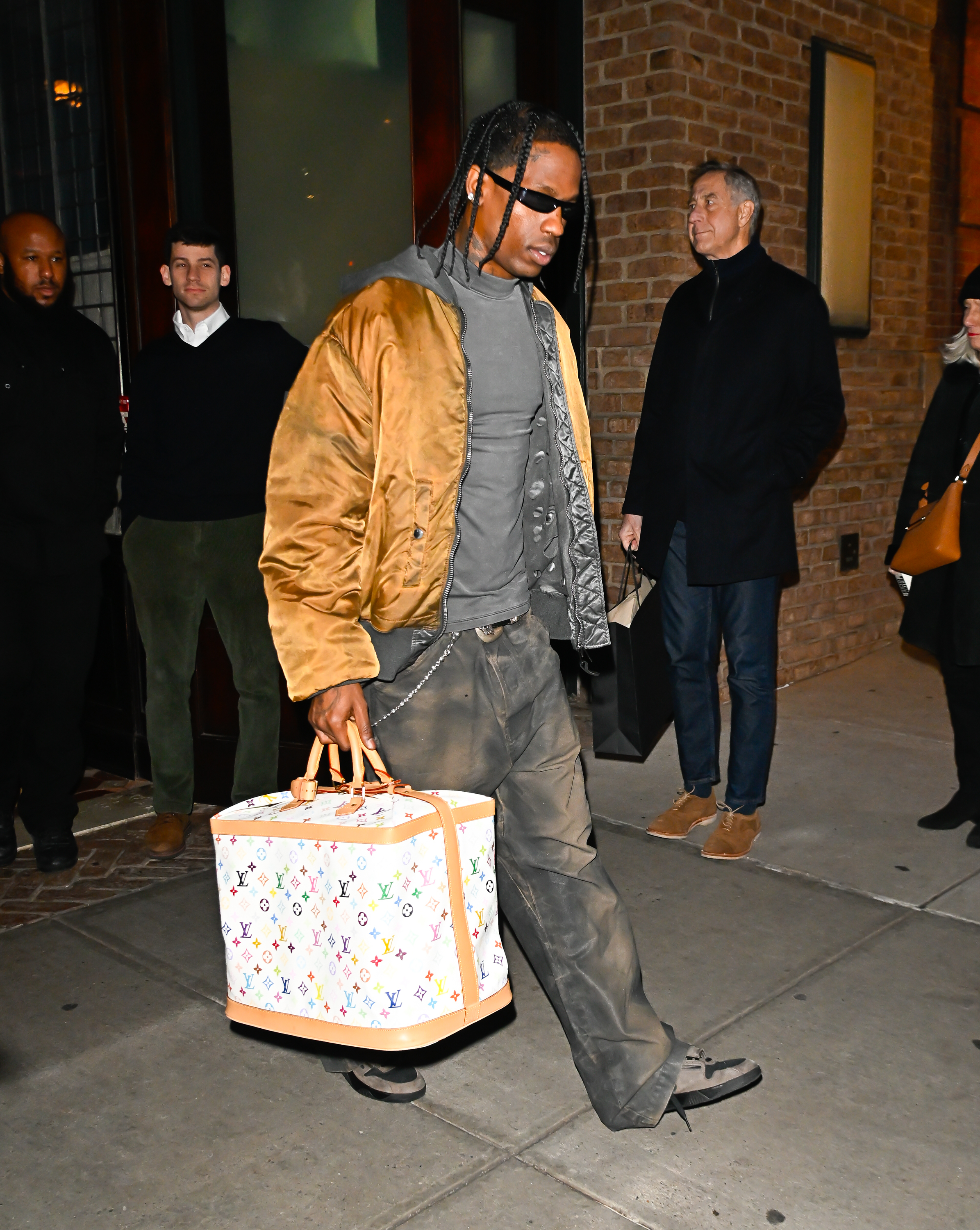 Travis Scott walks on a sidewalk carrying a large Louis Vuitton bag. He is wearing a bomber jacket, shirt, and dark pants with sunglasses