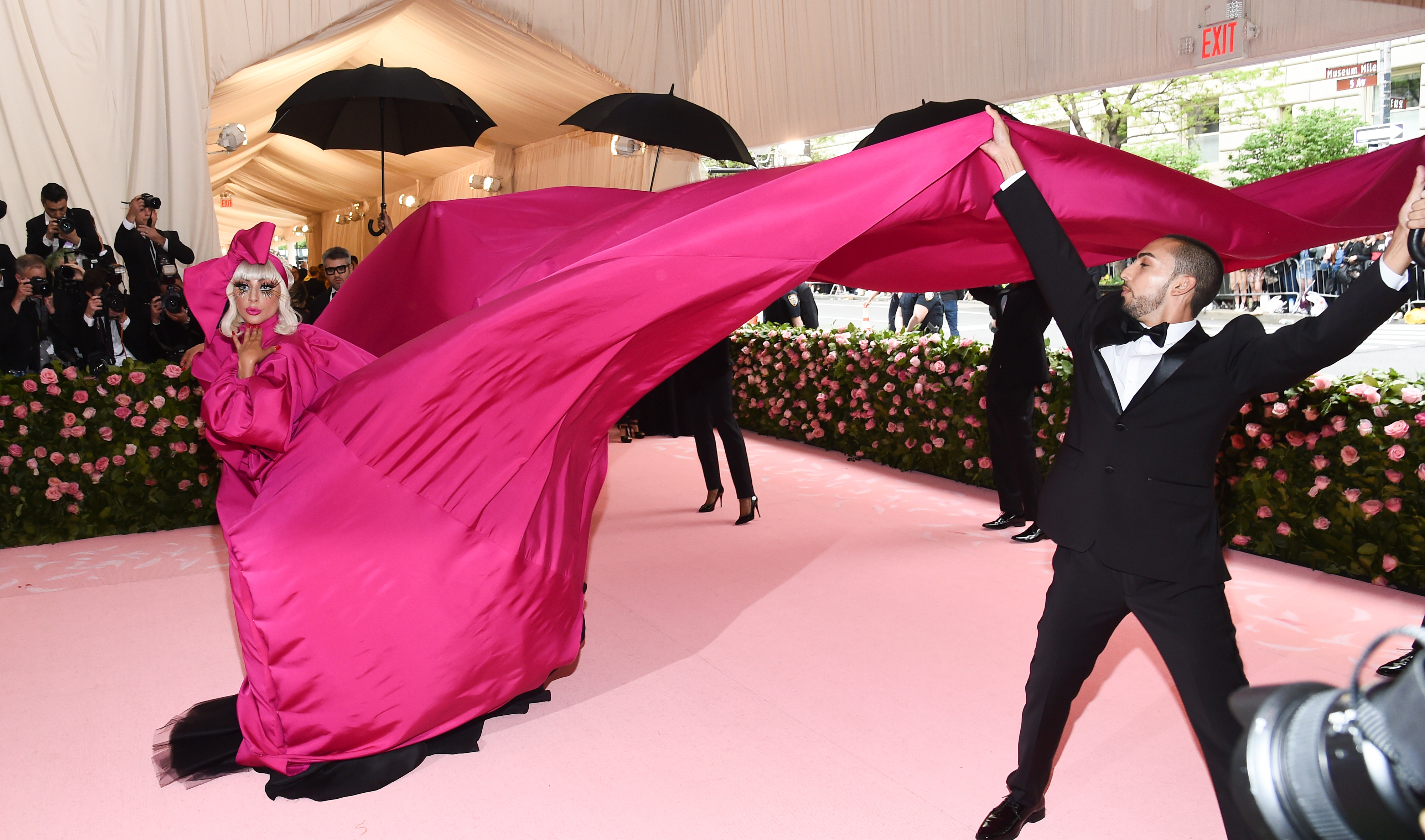 Lady Gaga in a dramatic floor-length pink gown with a large train, assisted by a man in a tuxedo holding part of the train