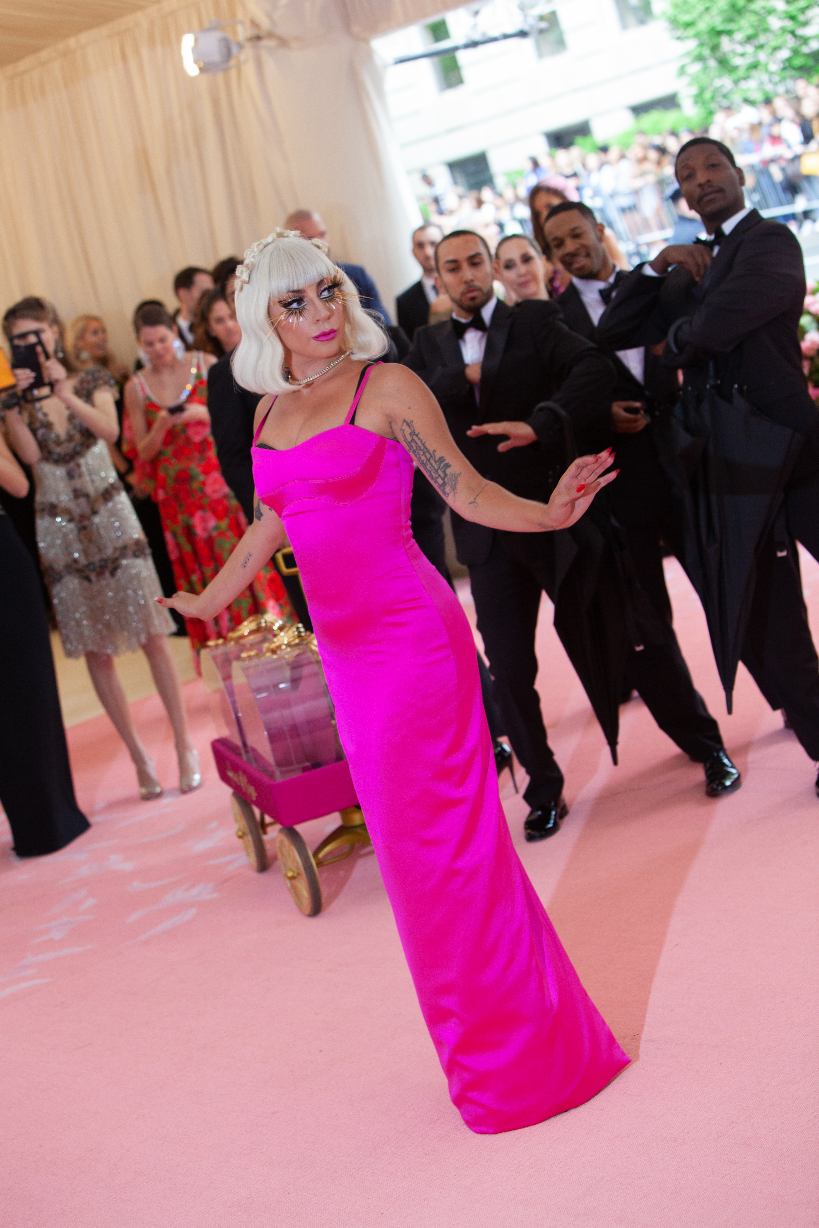 Lady Gaga is posing in a sleek column gown on the red carpet, with photographers and attendees in the background capturing the moment