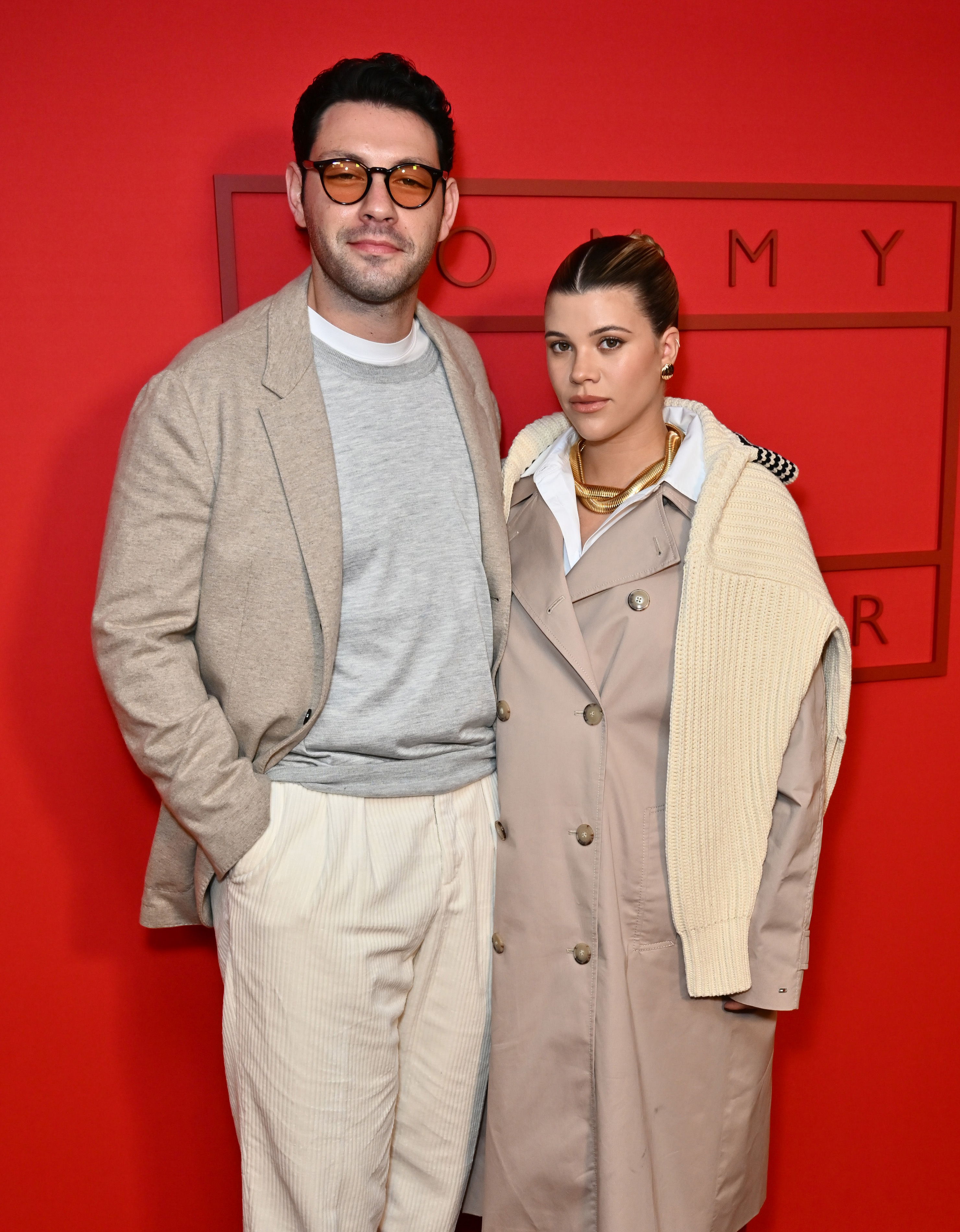 Elliot Grainge and Sofia Richie side by side at a Tommy Hilfiger event. Elliot wears a casual suit, and Sofia wears a trench coat and sweater over her shoulders