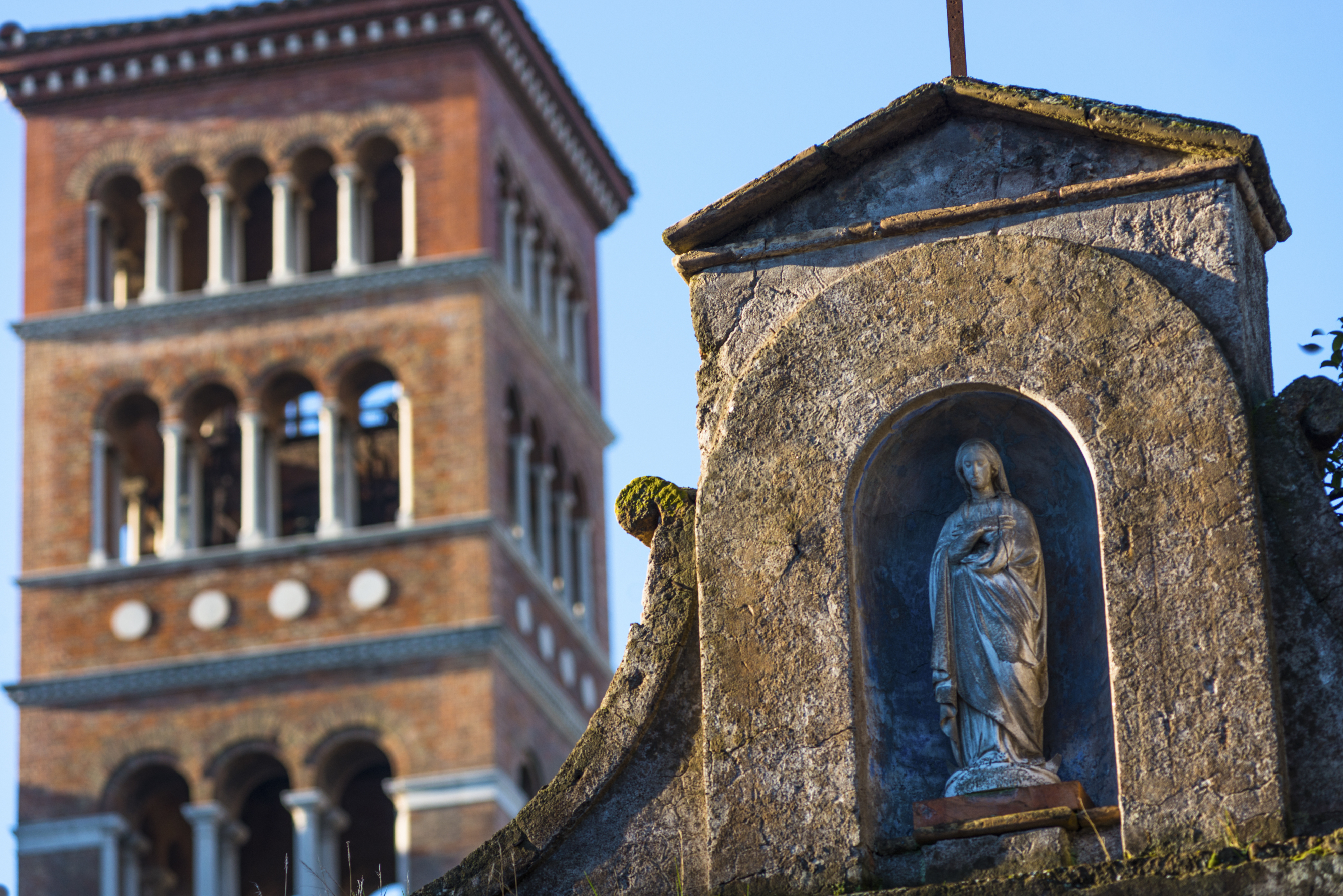 Statue of the Virgin Mary in an alcove of a stone structure with an Italian bell tower in the background