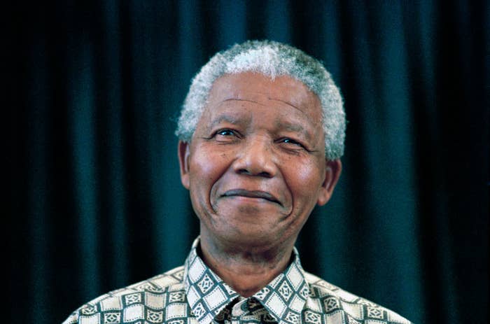Nelson Mandela dressed in a patterned shirt, smiling gently with a dark curtain backdrop