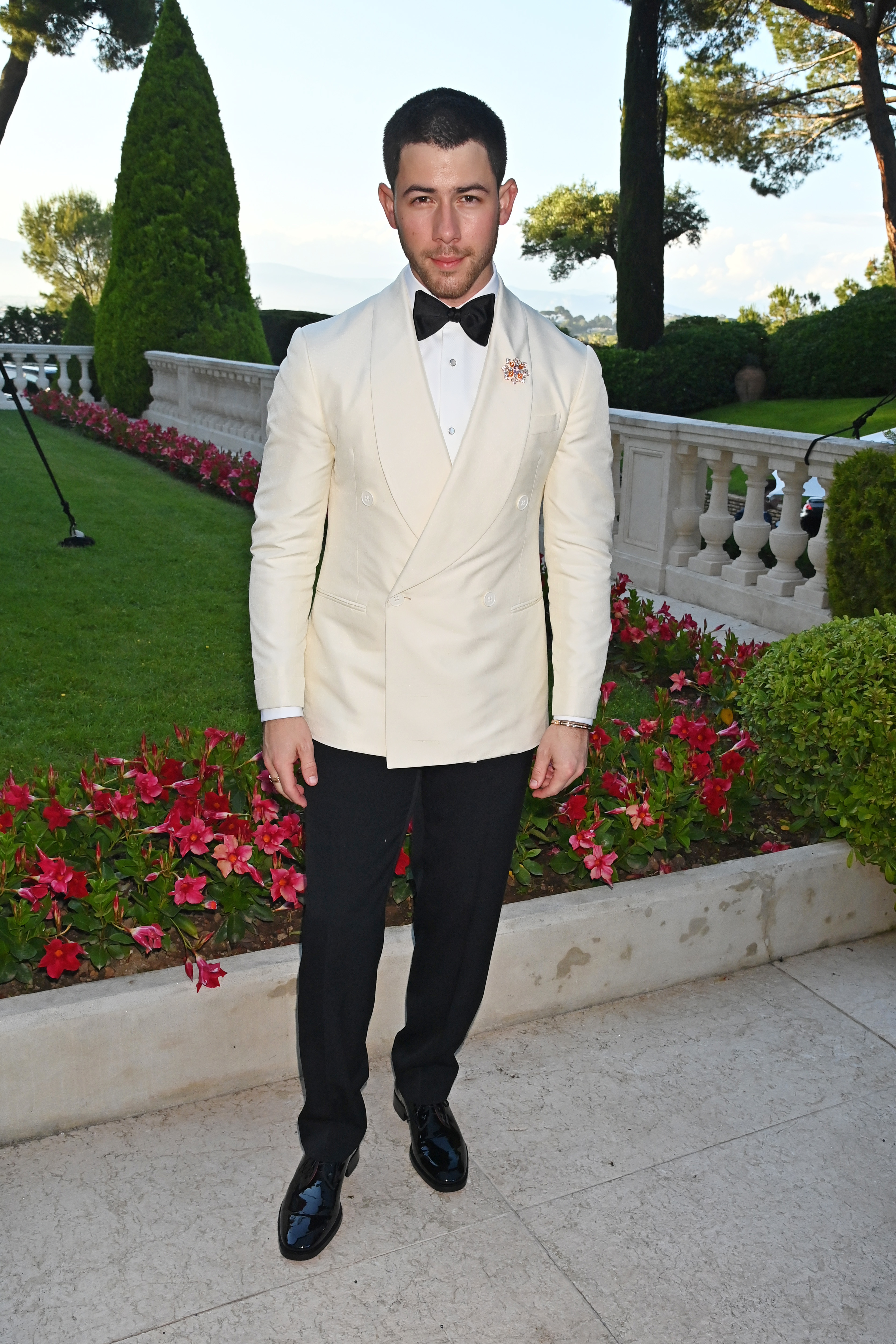 Nick Jonas stands outdoors, wearing a double-breasted cream tuxedo jacket with black pants and a bow tie. He poses in front of a garden with red flowers