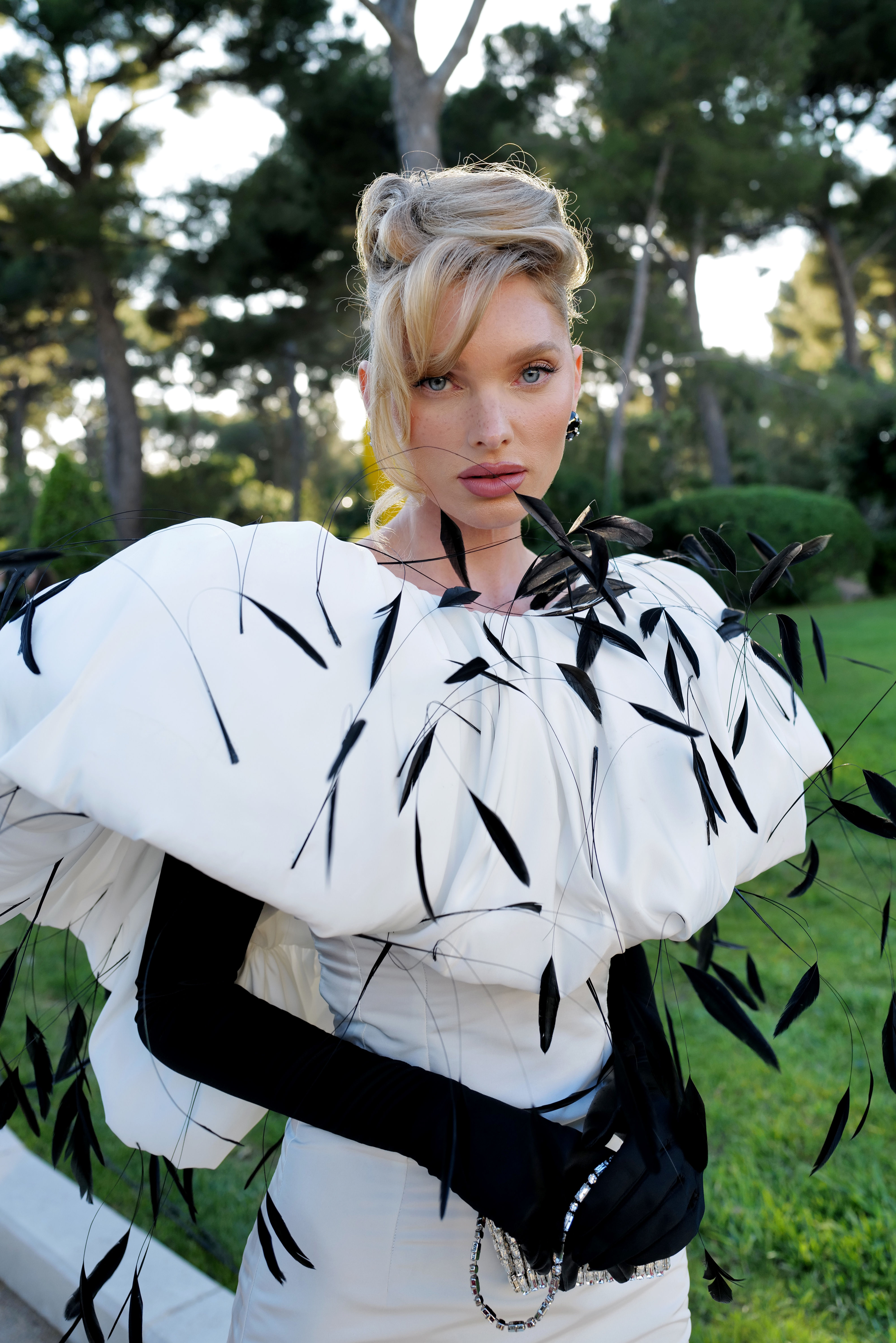 Elsa Hosk on a green lawn, wearing an elegant white dress with black accents and long black gloves, striking a pose