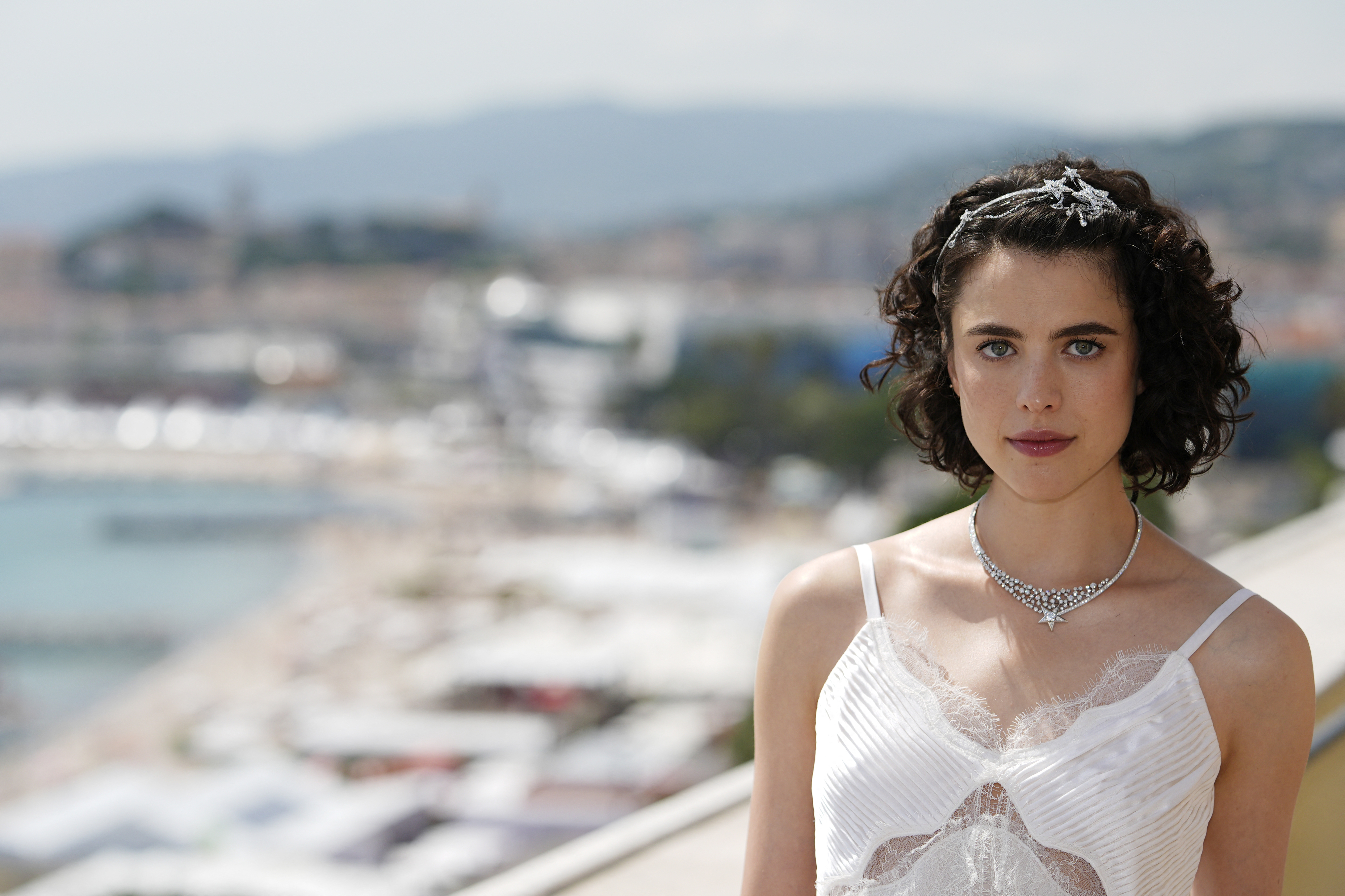 Margaret Qualley stands on a terrace overlooking a cityscape with a beach, wearing a white lace dress and a silver necklace