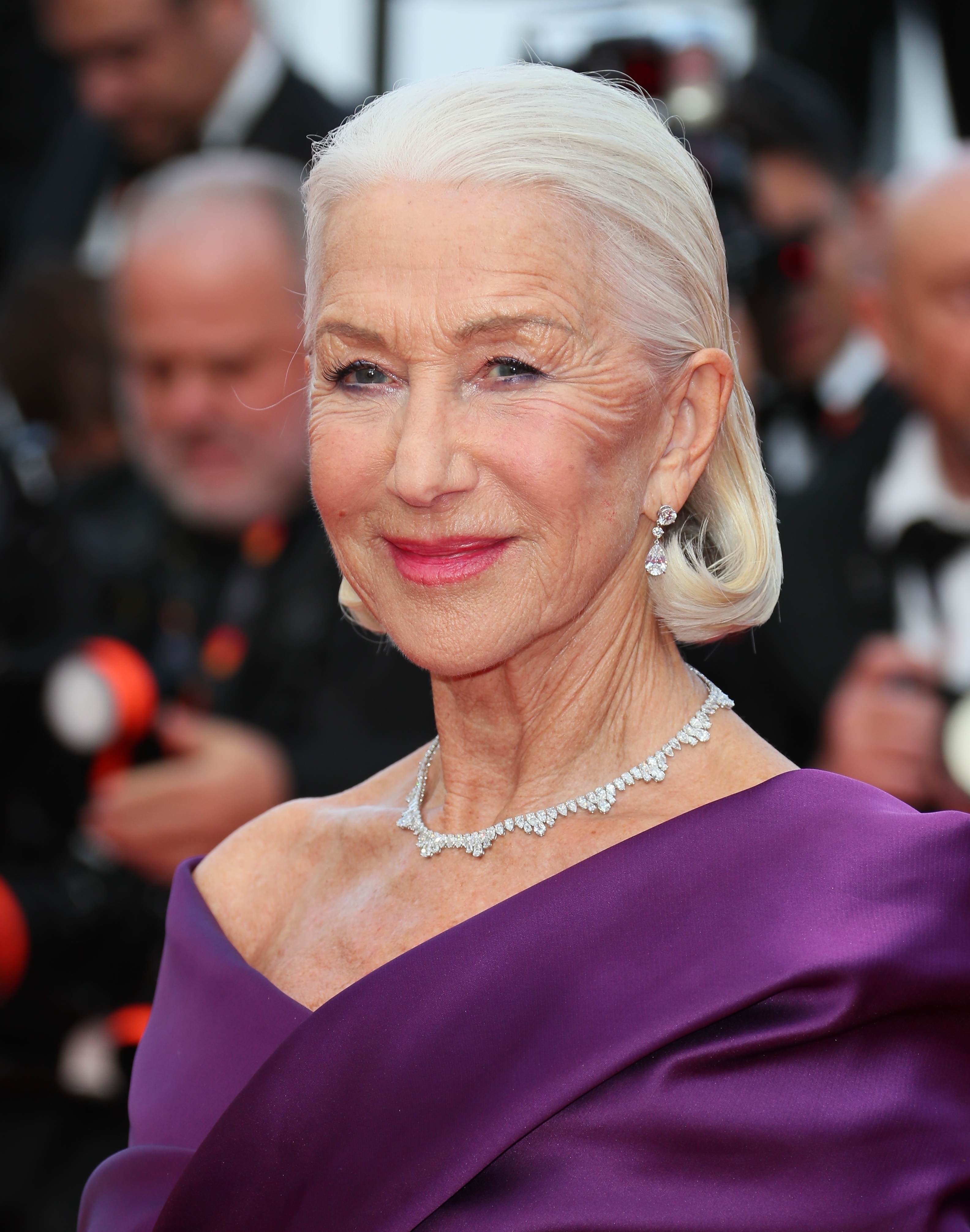 Helen Mirren poses on the red carpet in a chic, off-the-shoulder purple gown with elegant diamond jewelry, including earrings and a necklace
