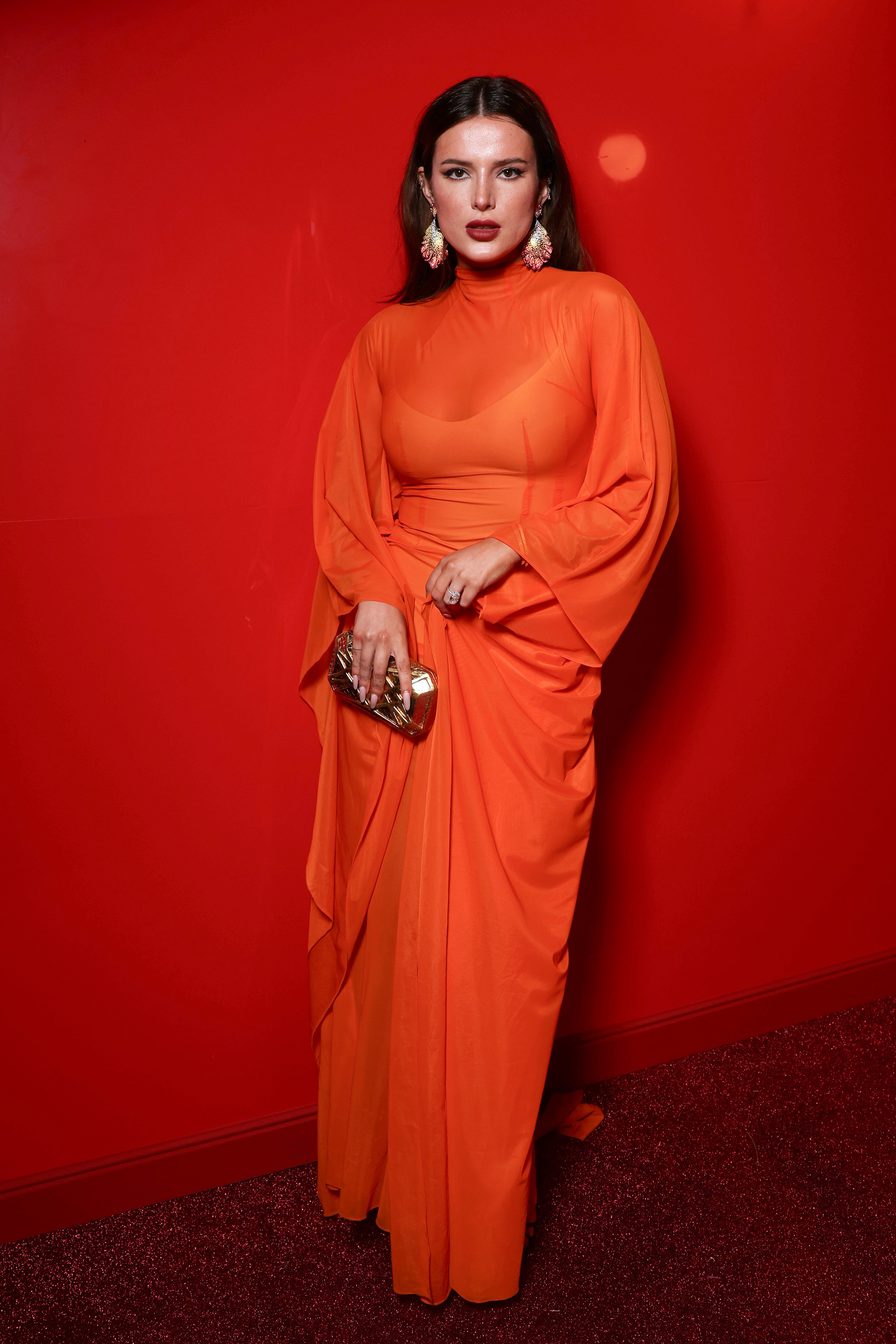 Bella Thorne in a long, flowing dress with loose sleeves, holding a metallic clutch and wearing statement earrings, posing against a plain background