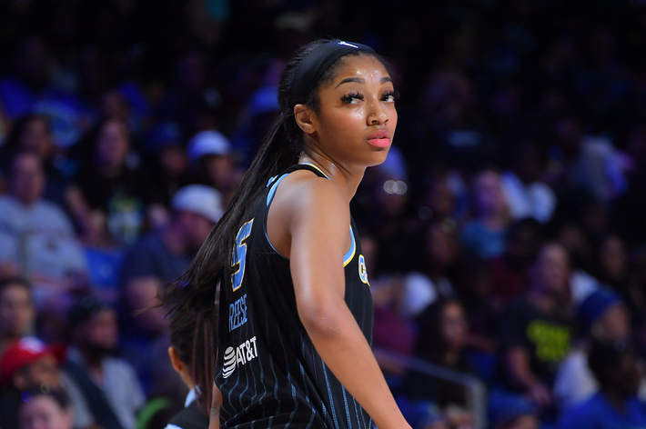 WNBA player A'ja Wilson looks back over her shoulder on the basketball court during a game, wearing her team jersey and shorts. Fans are visible in the background