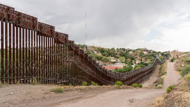 A section of the US-Mexico border wall in a rural area, featuring barbed wire and a dirt road running alongside it