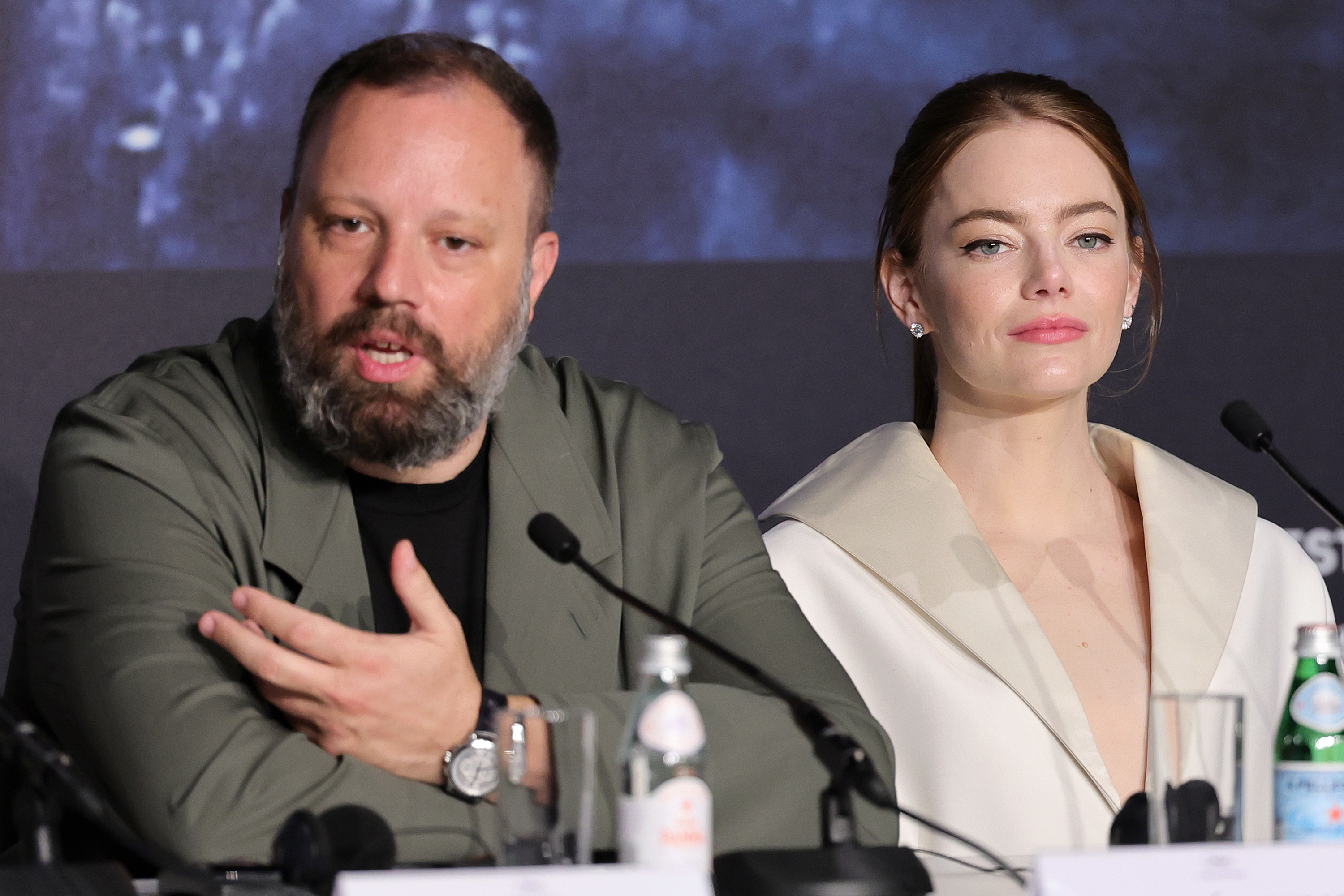 Yorgos Lanthimos speaking at a press event, with Emma Stone sitting beside him in a press conference setting