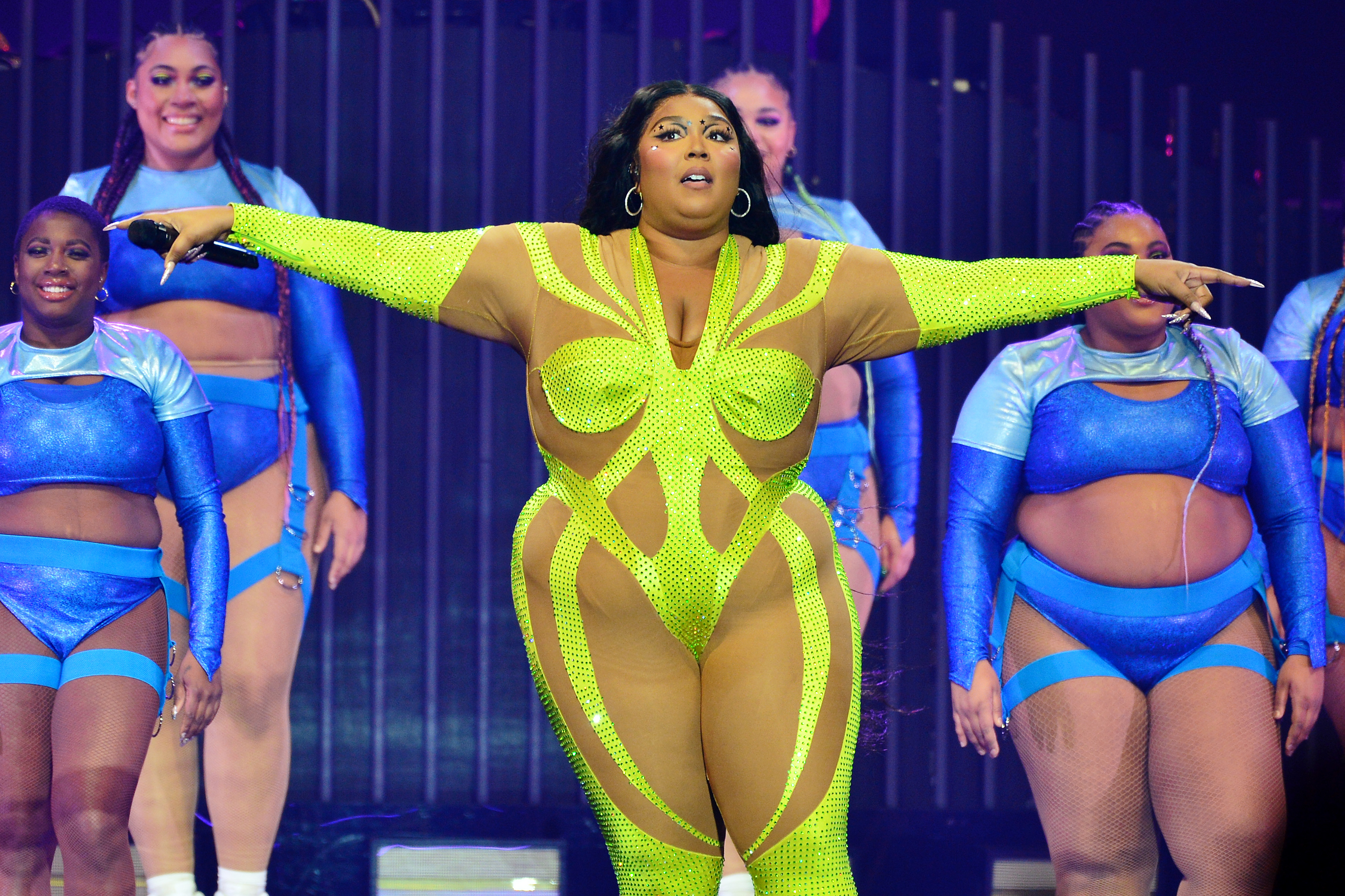 Lizzo performs on stage in a bright, futuristic outfit with intricate cutouts, surrounded by backup dancers in coordinated costumes