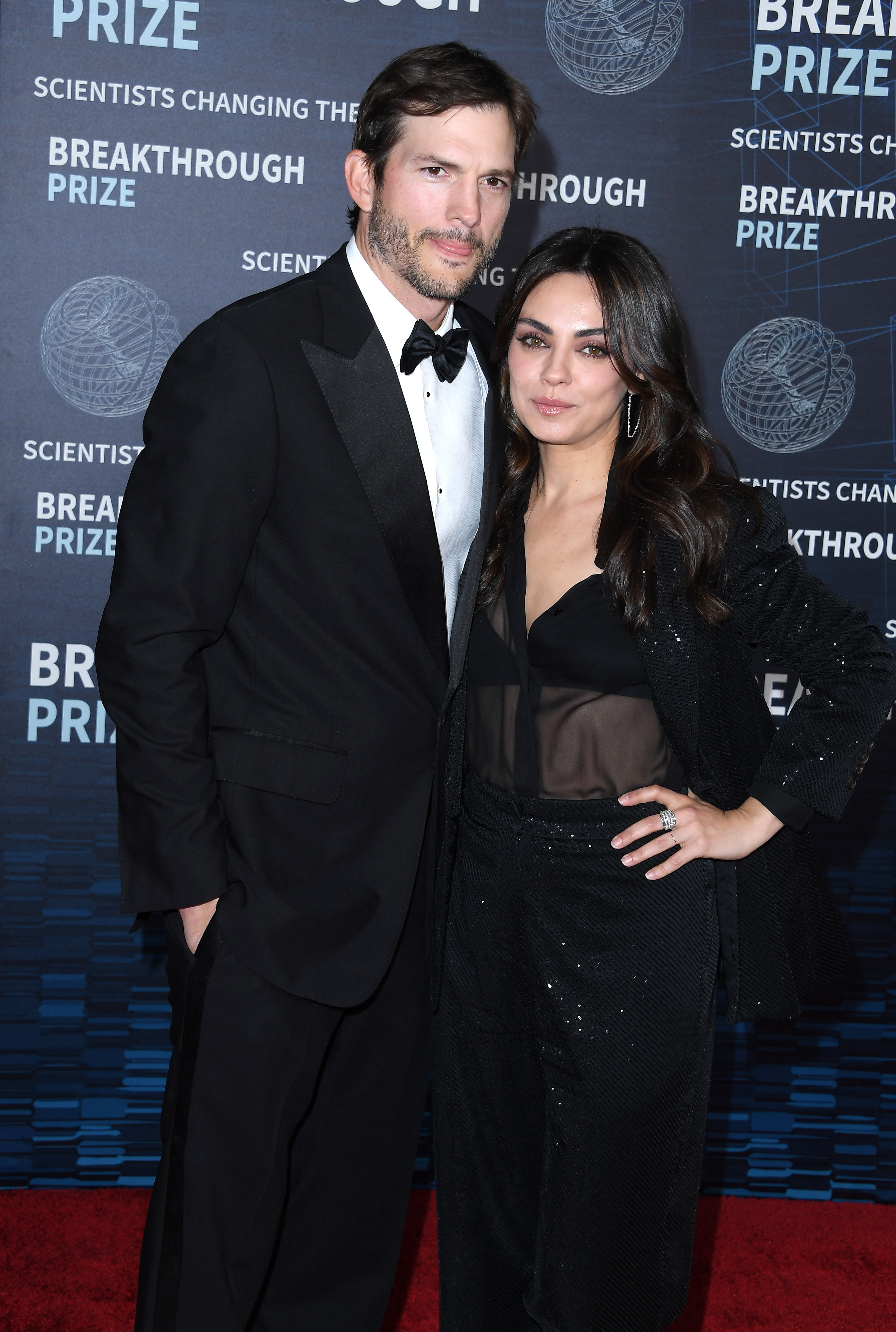 Ashton Kutcher in a black tuxedo and Mila Kunis in a sheer top and black pants at the Breakthrough Prize red carpet event