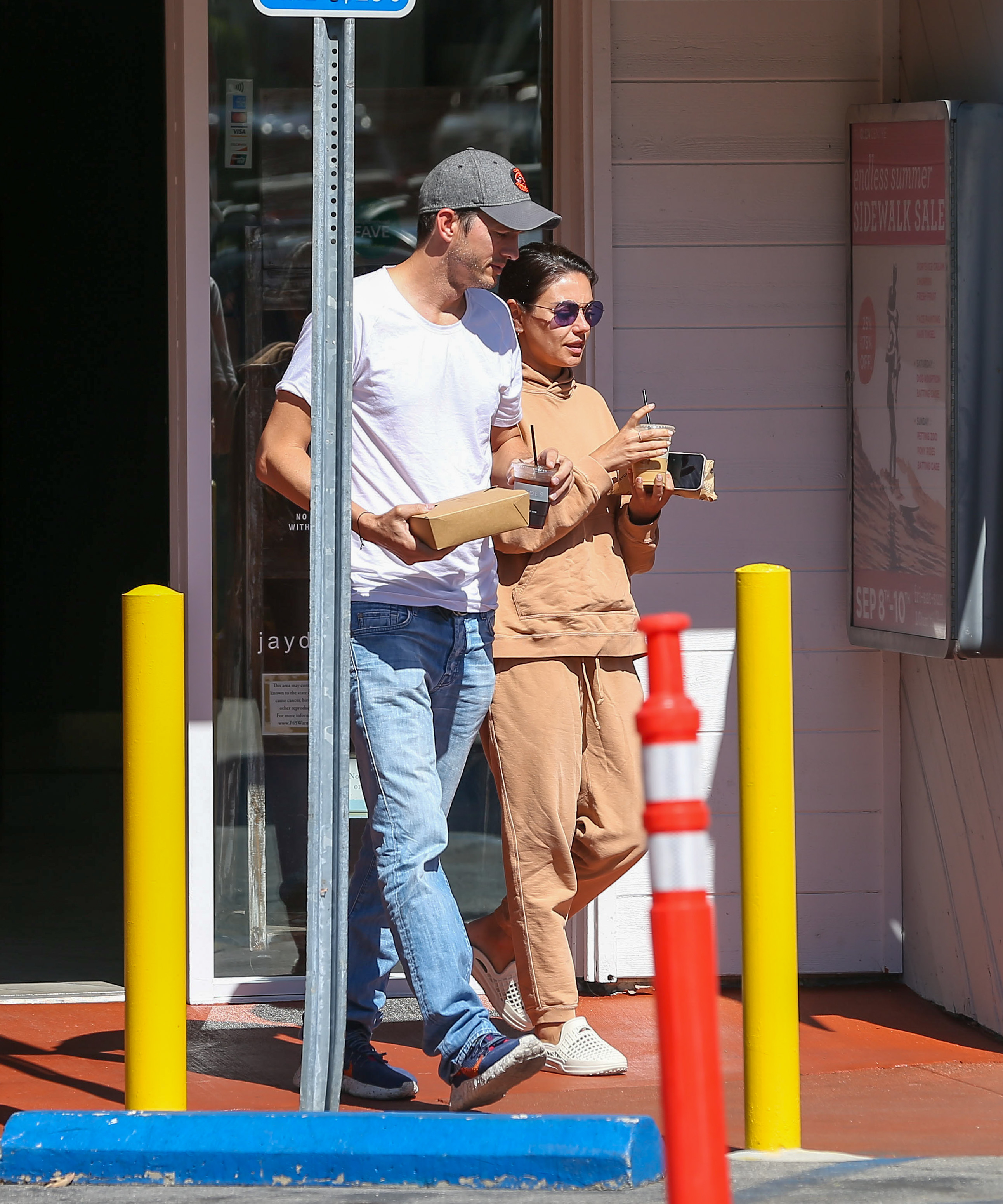 Ashton Kutcher and Mila Kunis are casually dressed and holding drinks and food containers while walking outside a building